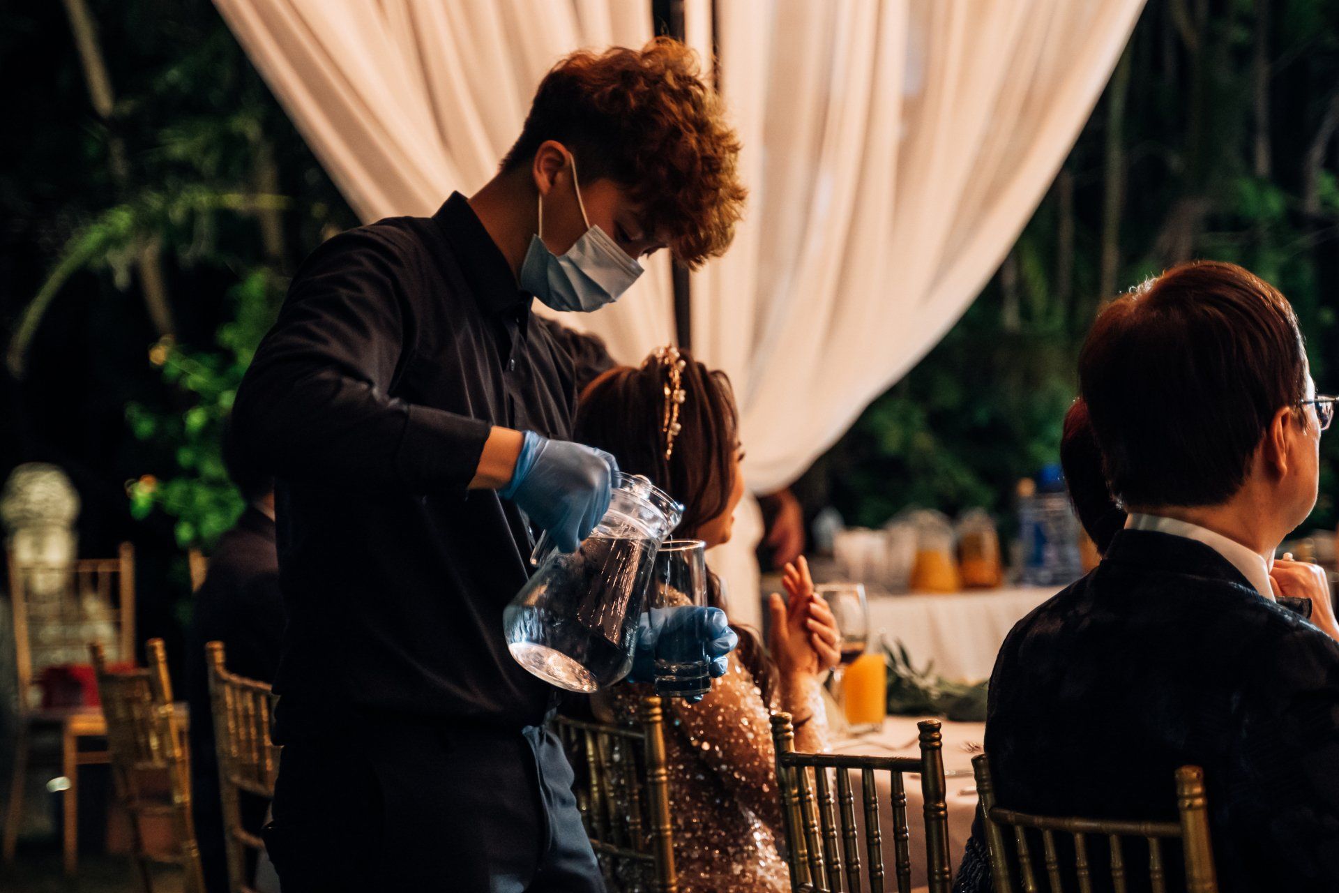 Waiter pouring water into a glass at a table in an outdoor event. He wears a mask and gloves.