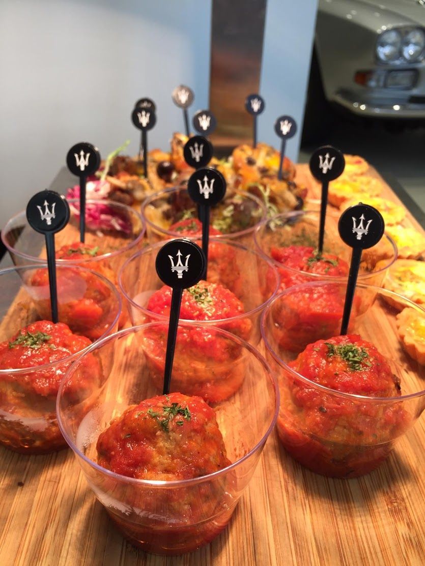 Appetizers in clear cups topped with tomato sauce and a trident pick, on a wooden board.