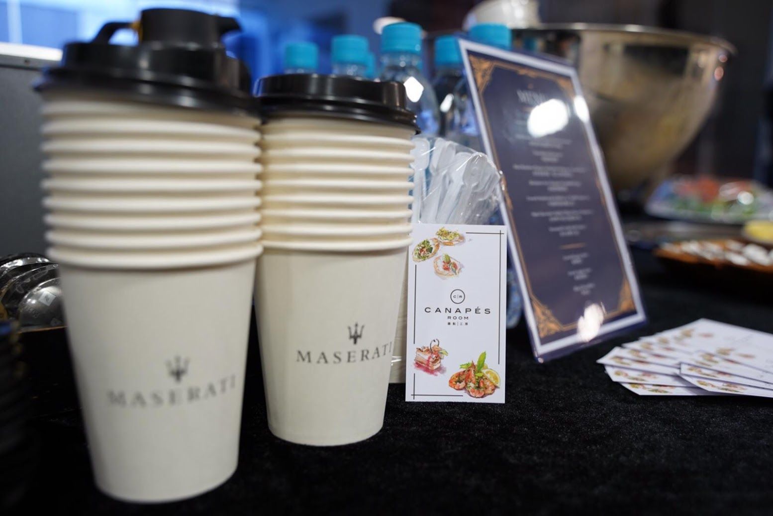 Two Maserati branded coffee cups, menu, and water bottles on a table.