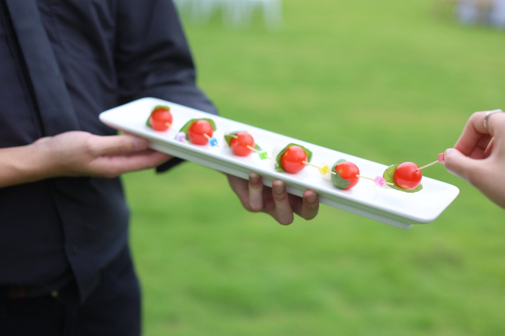 Server holding tray of cherry tomatoes on skewers, offering one to a person outdoors.