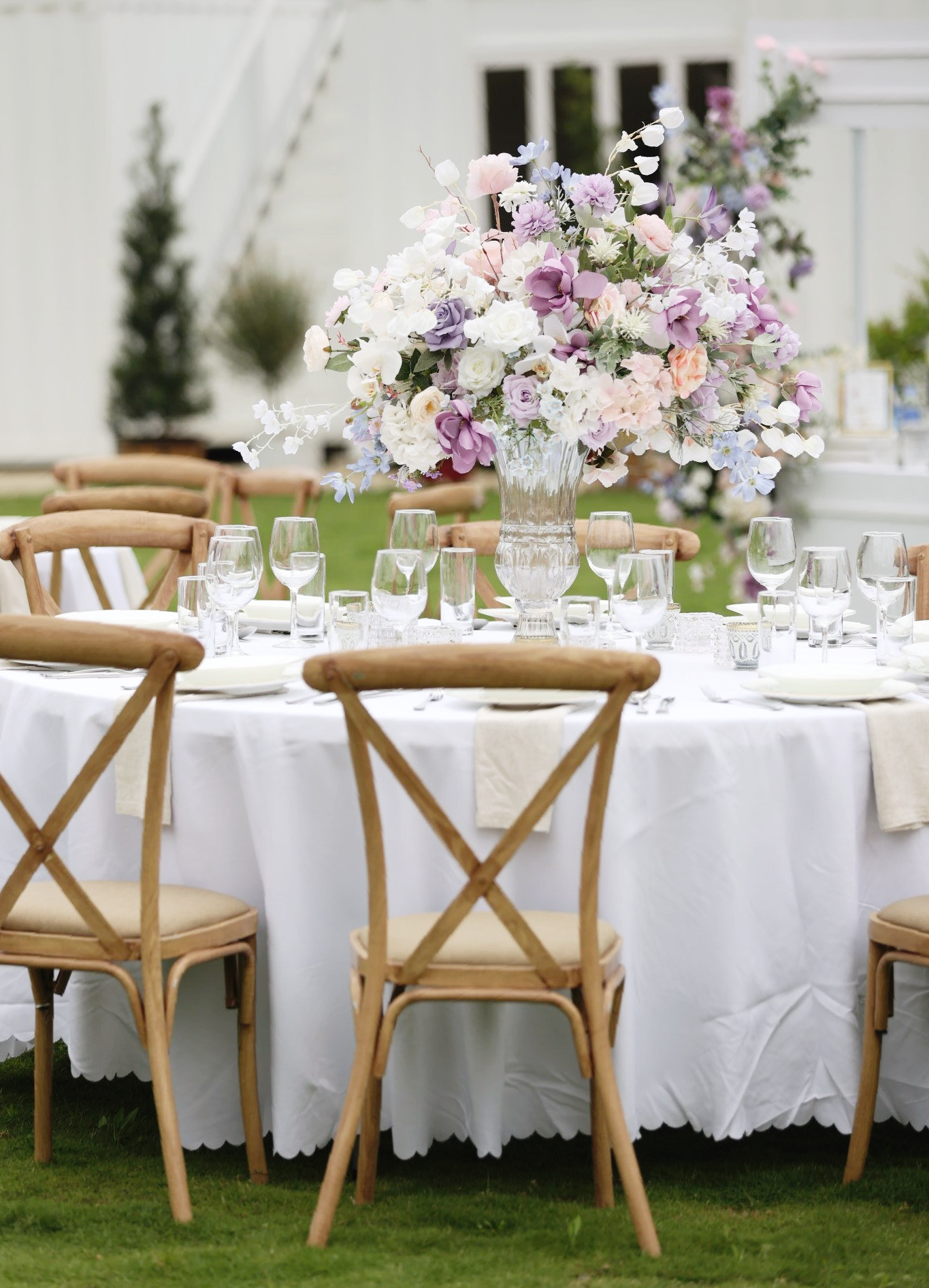 Round table set for a formal outdoor event, with floral centerpiece and cross-back chairs.