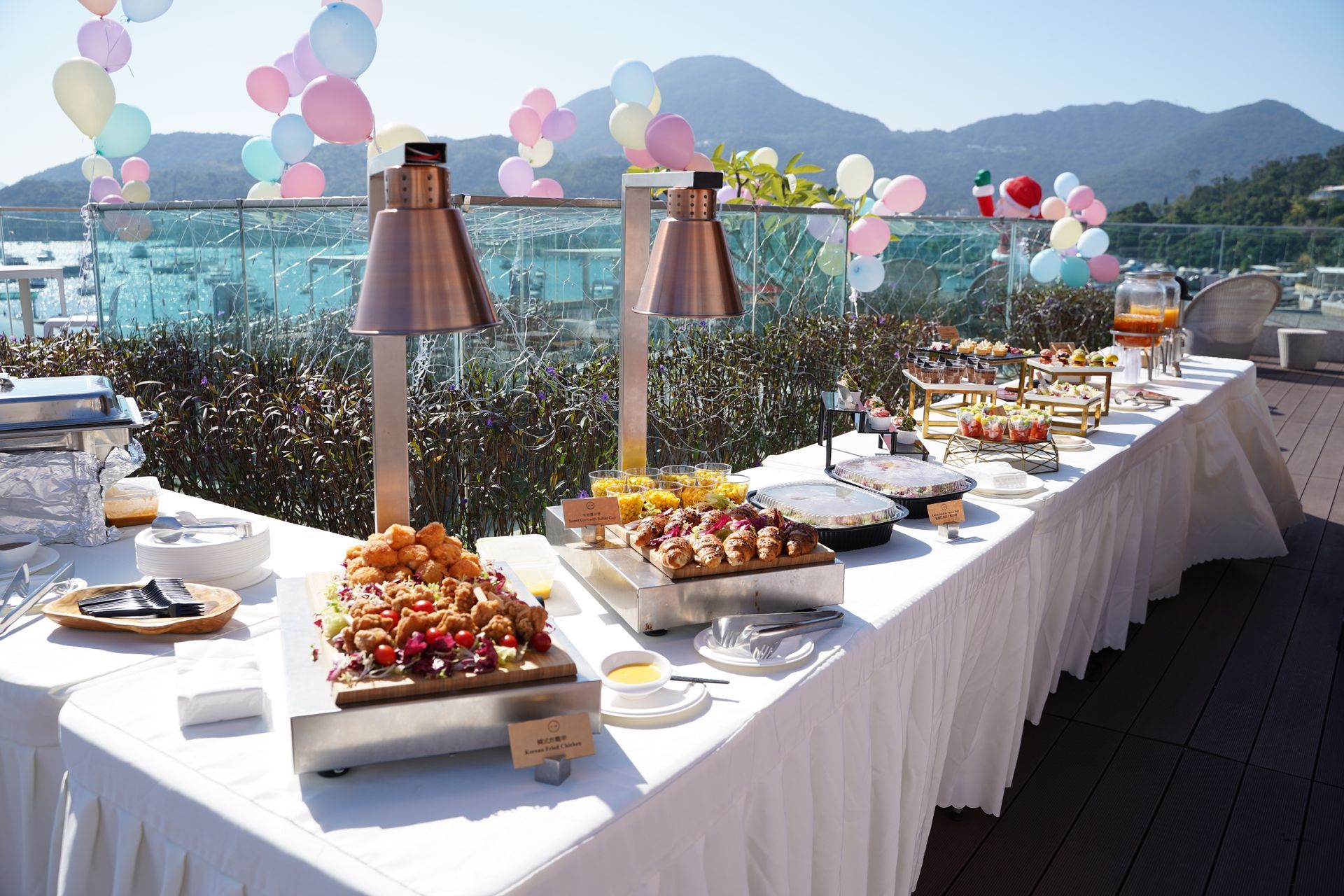 Outdoor buffet with food under heat lamps. Balloons and mountains in the background.