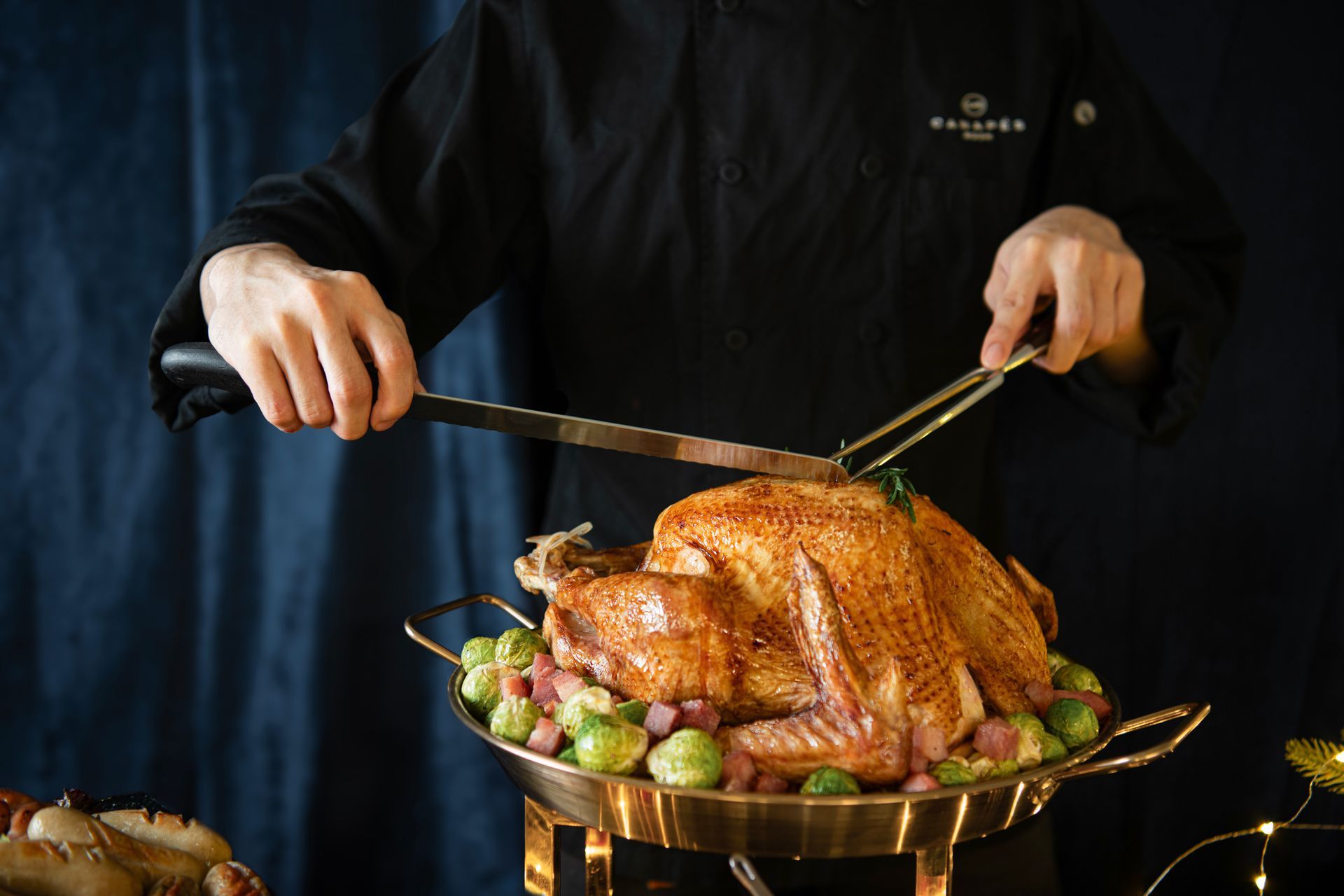 Chef carving a roasted turkey, set on a bed of Brussels sprouts and ham in a gold pan.