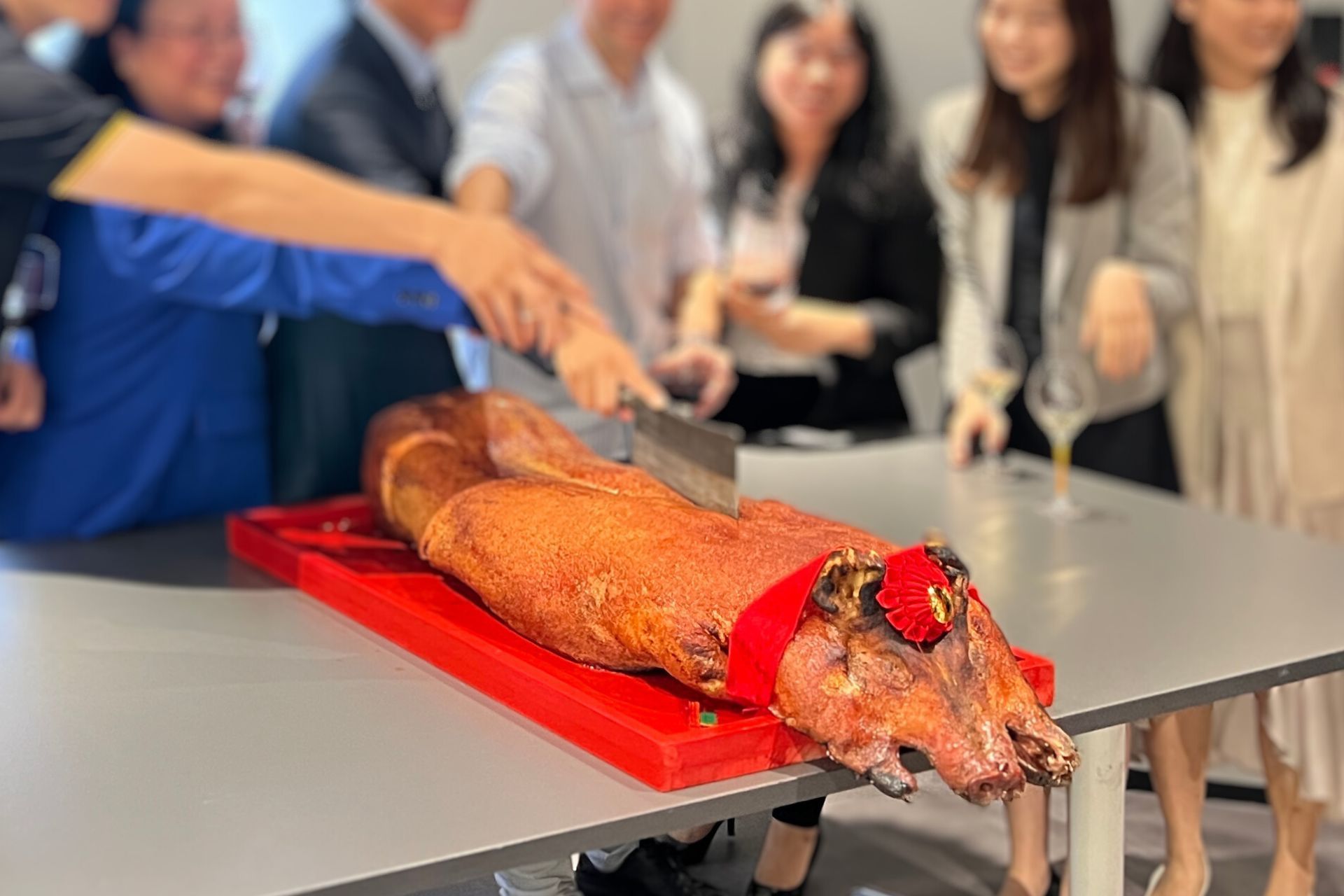 Group cuts a roasted pig on a table; red cutting board. People watch in an office setting.