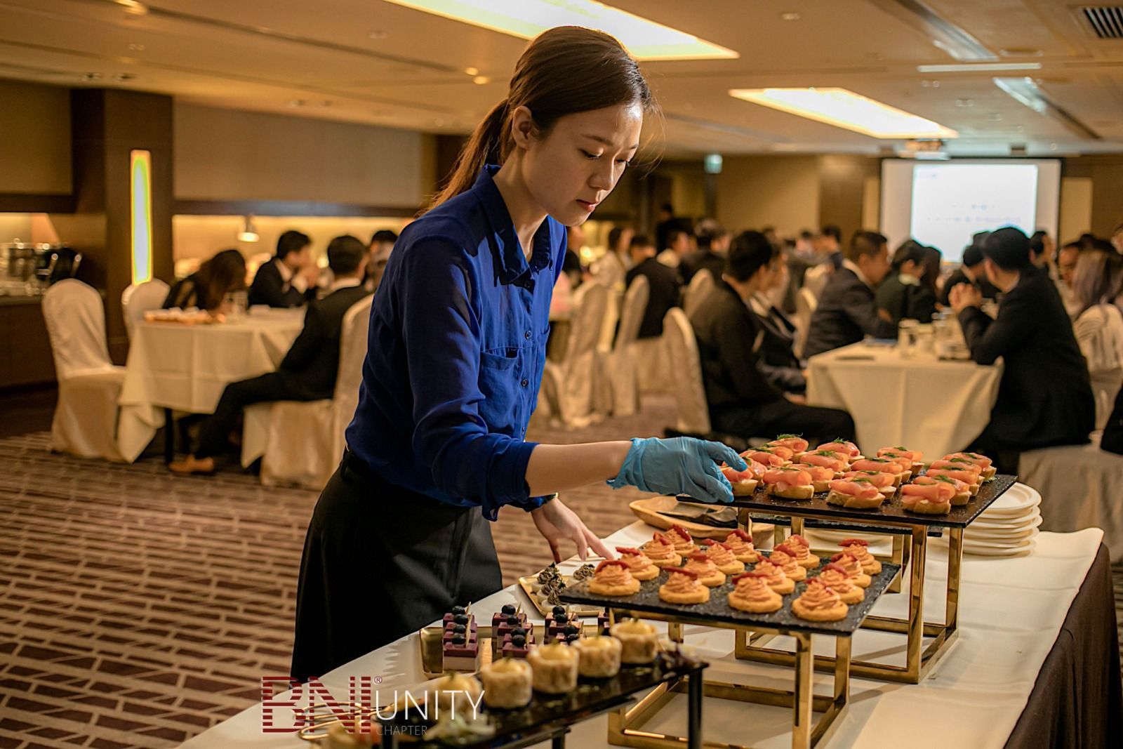 A server placing appetizers at a catered event. People seated at round tables in the background.