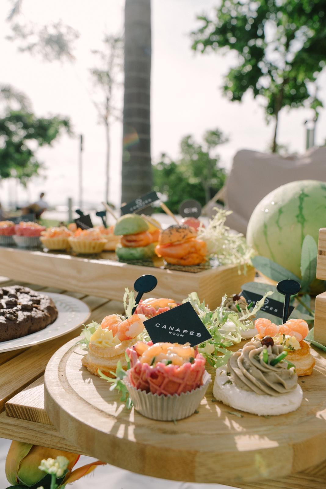 Outdoor dessert display: Mini tarts, waffles, watermelon, and appetizers on wooden platters. Sunny setting with palm trees.