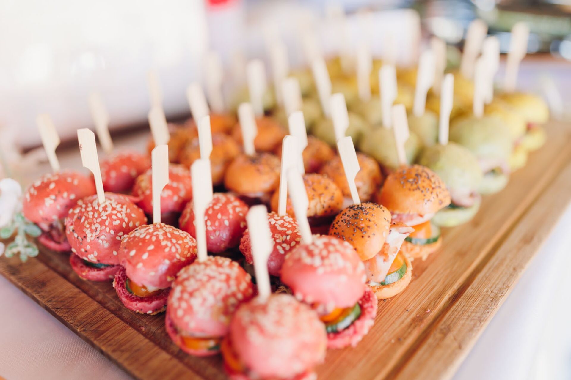 Miniature colorful burgers on wooden board, skewered, arranged in rainbow order.