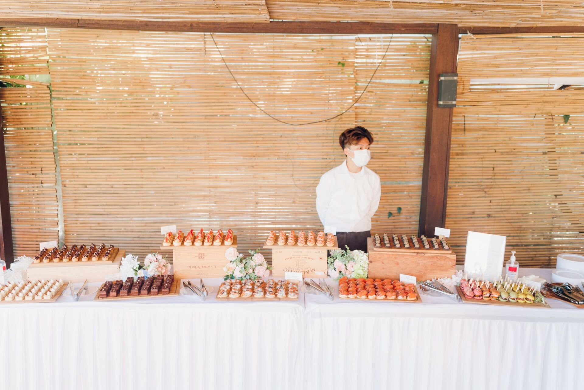 Person wearing mask stands behind a table laden with pastries, against a woven bamboo backdrop.