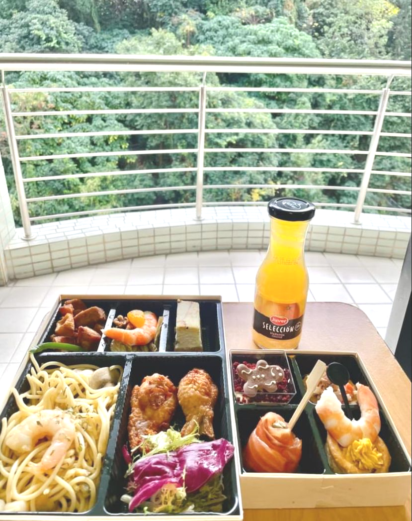 Bento box with various foods and orange juice bottle on a table with a balcony view of green trees.