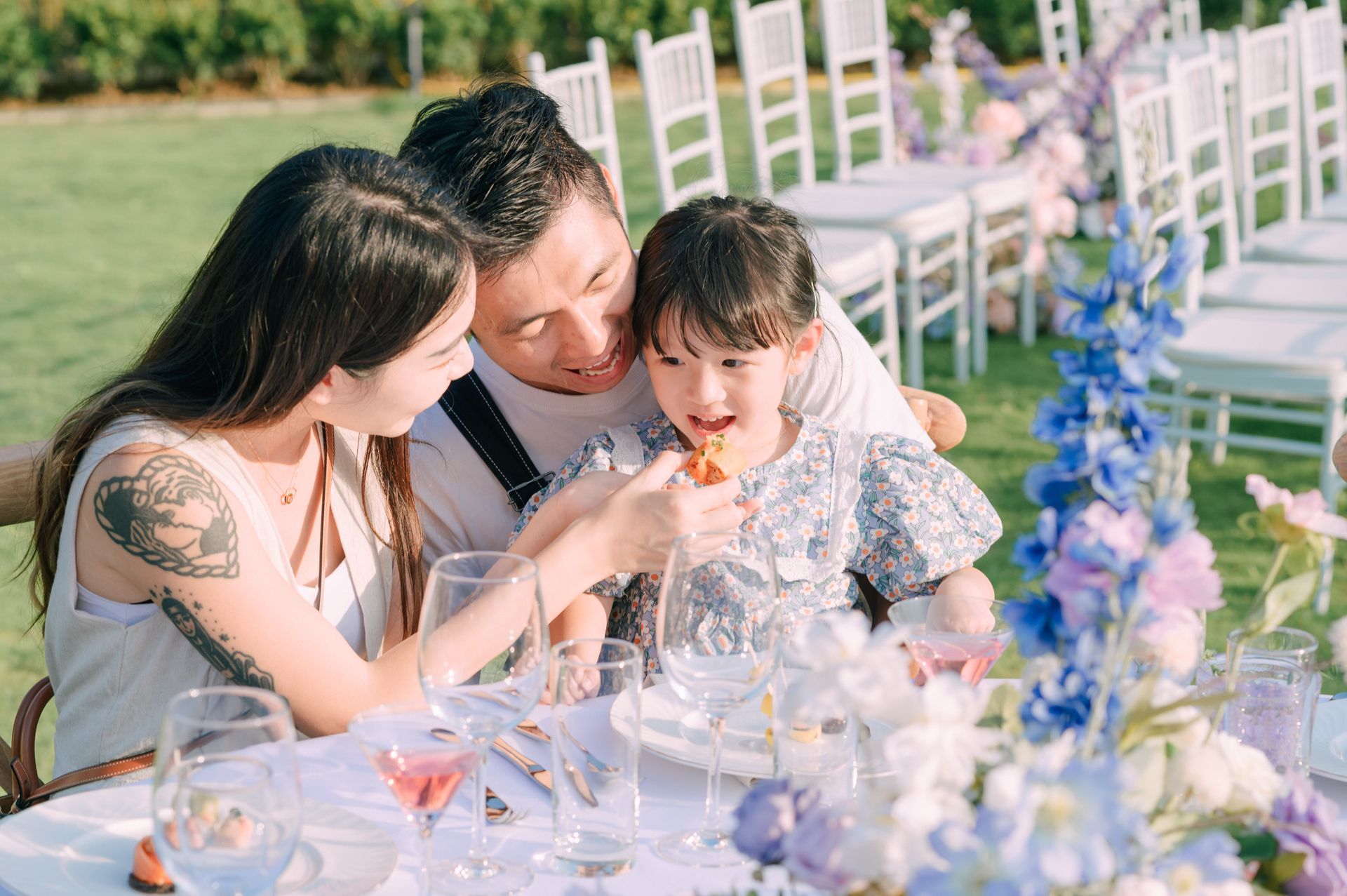 Family at a decorated outdoor table: parents feed a young child, floral arrangements in soft colors.