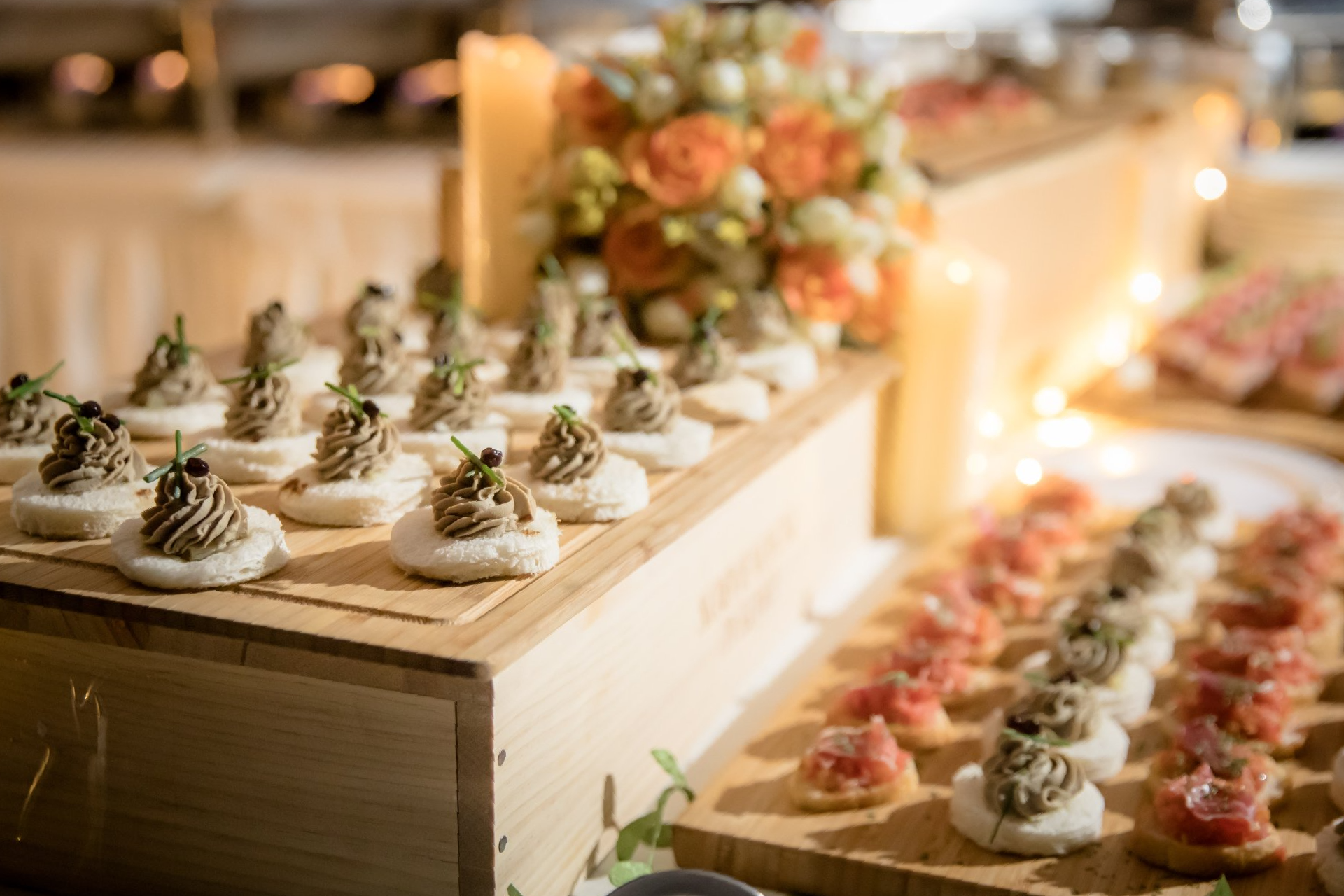 Appetizers on wooden display: bread topped with savory spread, floral arrangement and candles in background.