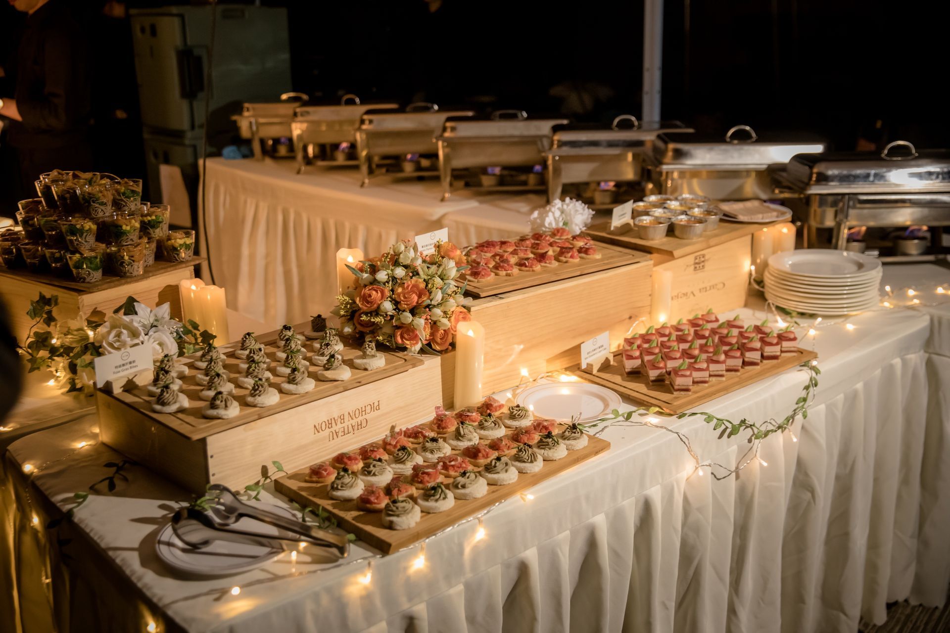 Buffet table with various appetizers and warmers, decorated with lights and flowers at an event.