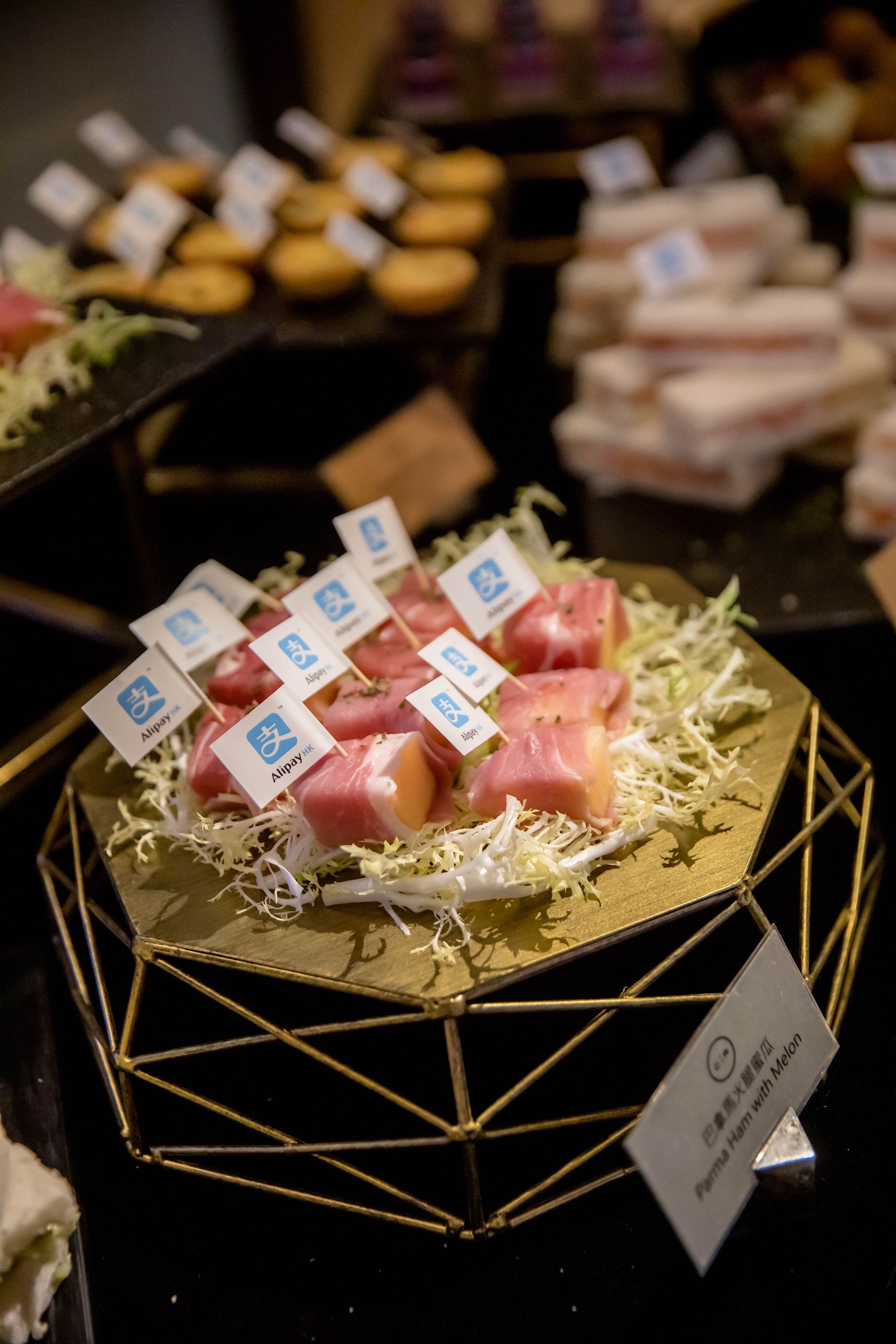 Appetizer display: prosciutto-wrapped bites on a gold geometric stand, topped with flags, other foods in background.