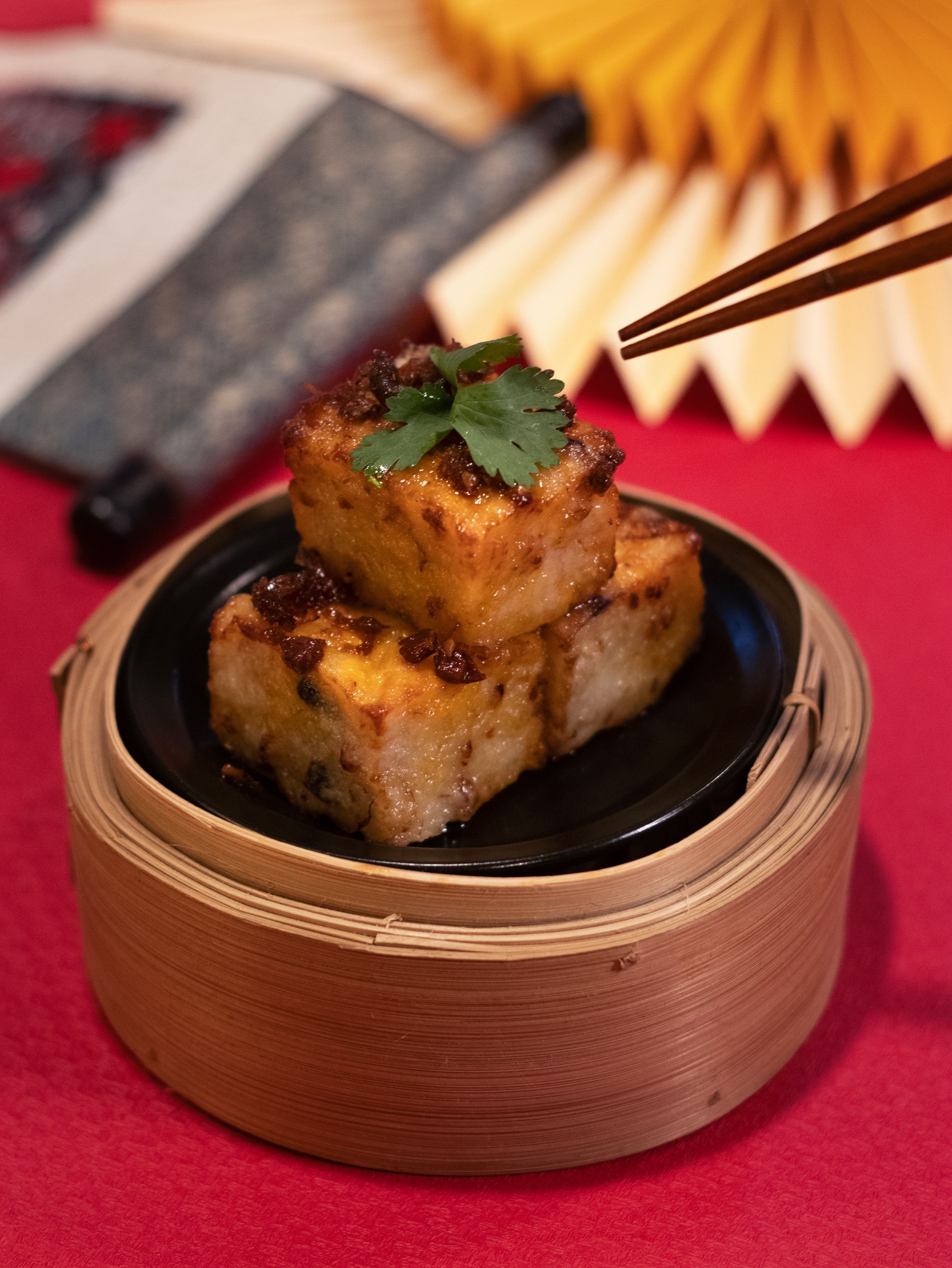 Tofu cubes in a bamboo steamer, topped with sauce and cilantro. Red background, chopsticks reaching in.