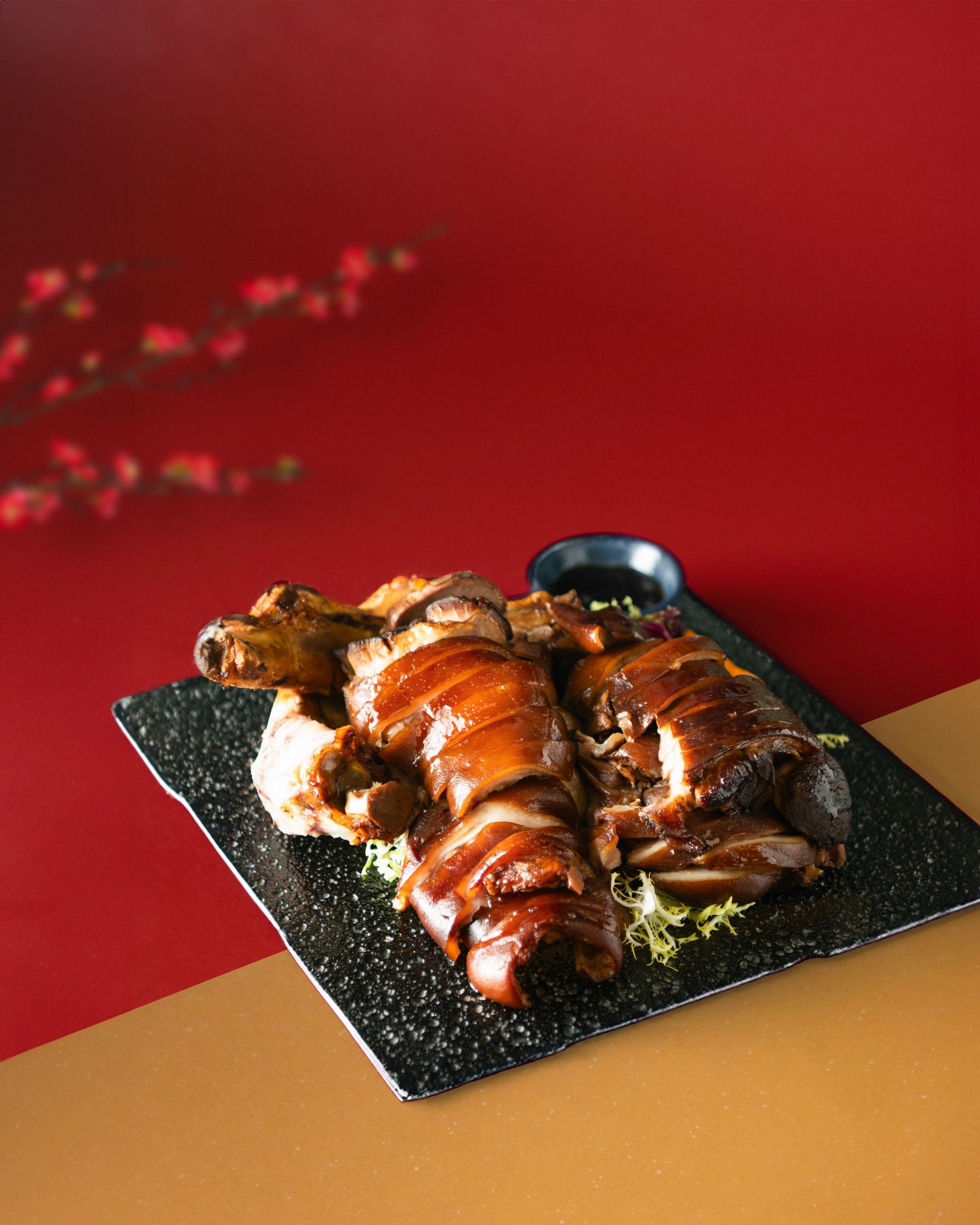 Braised pork trotters on a black plate with dipping sauce against a red and gold background.
