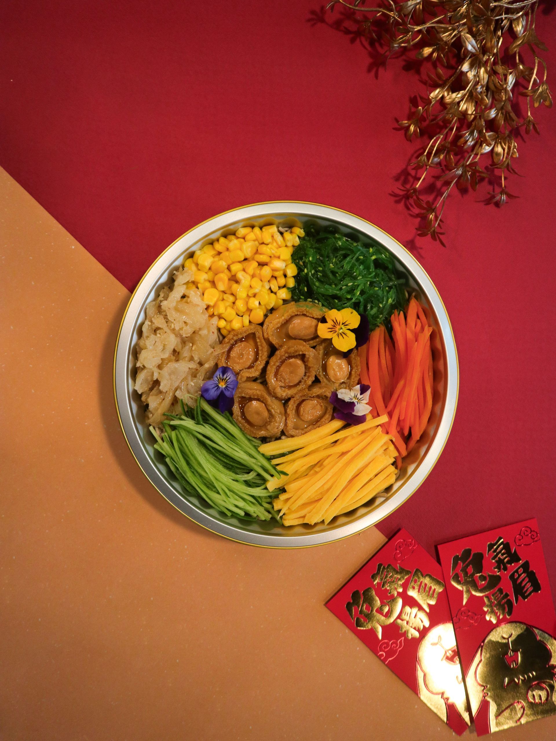 A platter of colorful, prepared food, including abalone, corn, and vegetables, on a red and tan background.