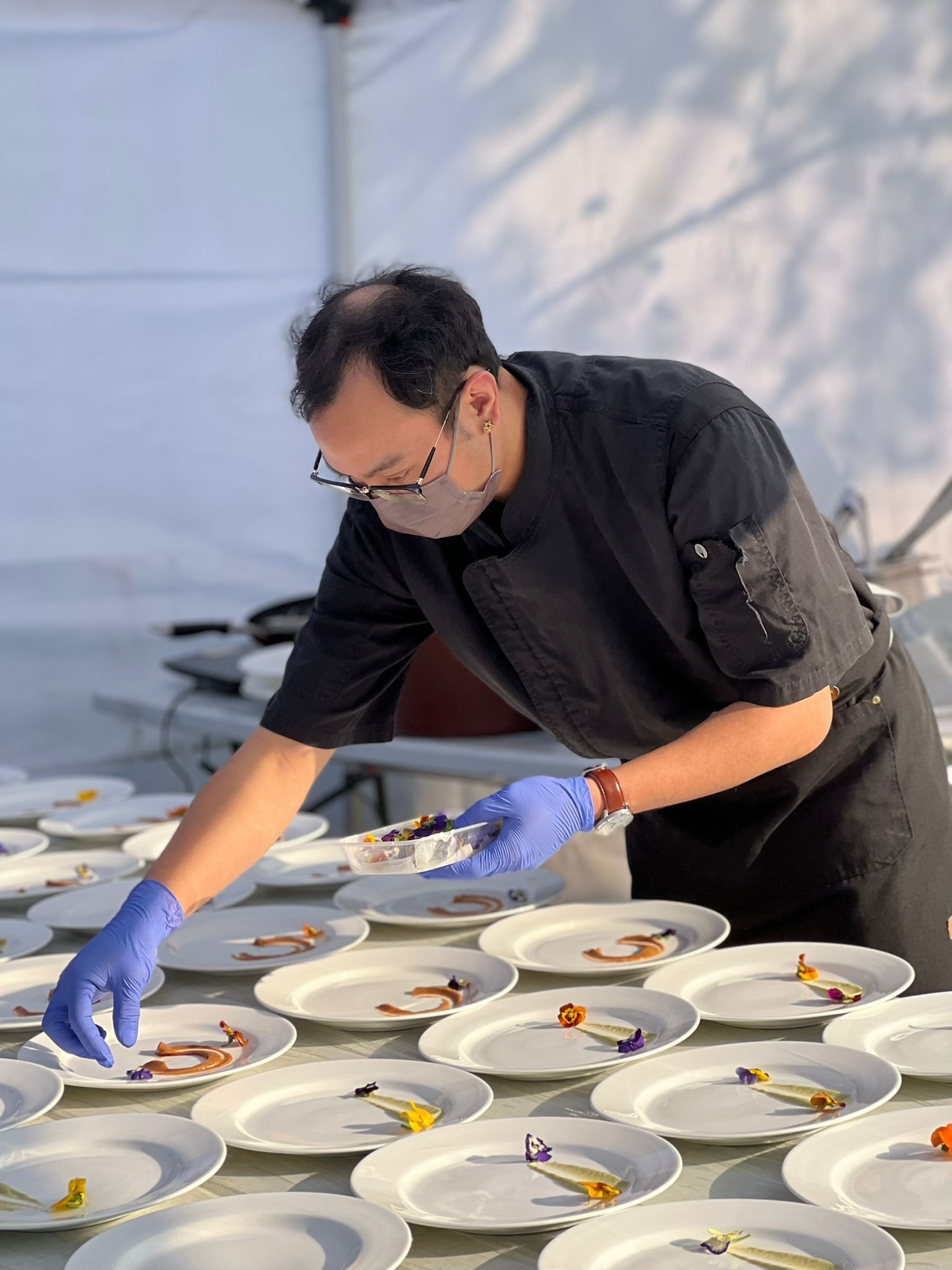 Chef in black uniform and face mask arranges food on plates, outdoors.