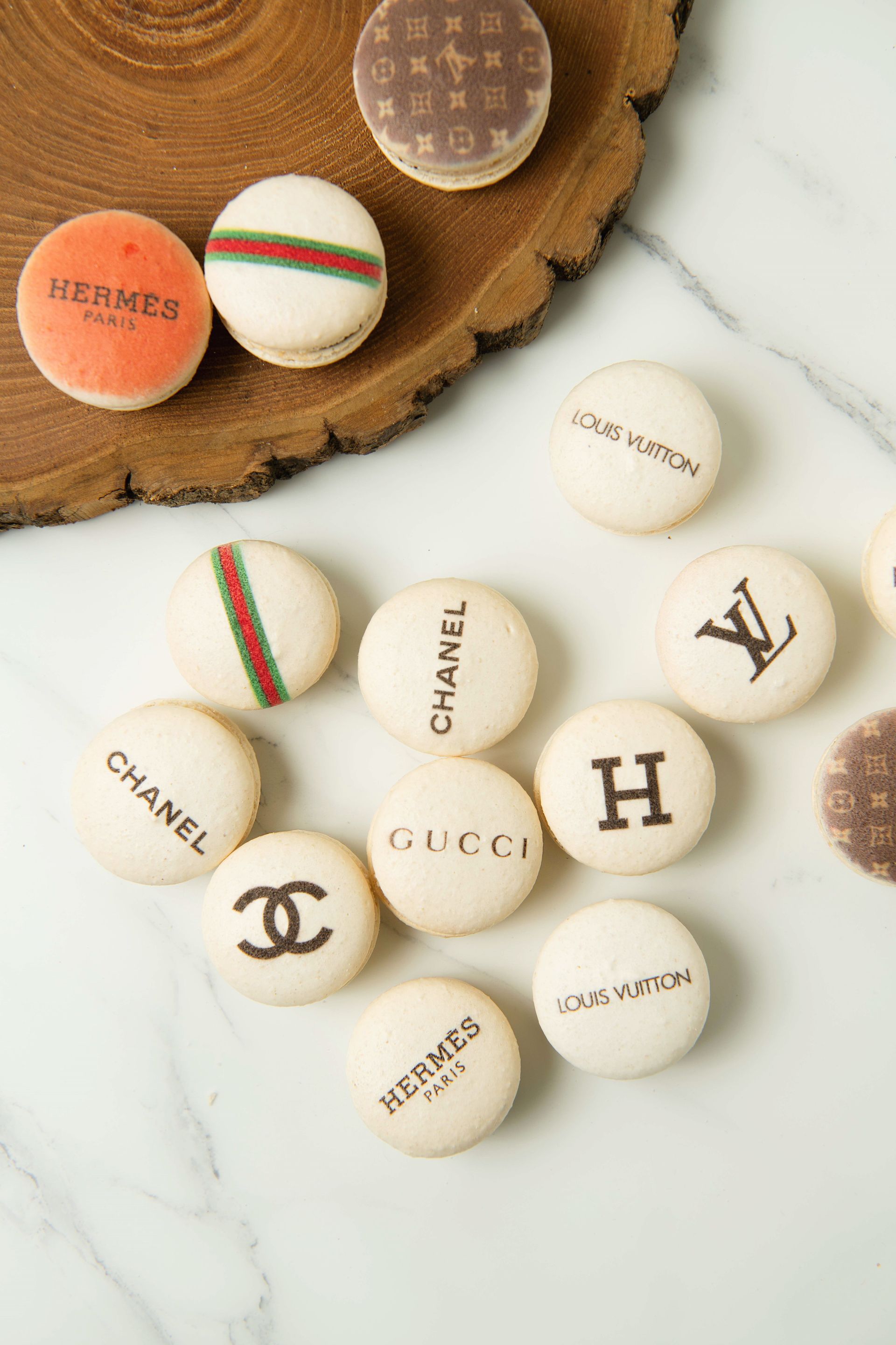 Macarons decorated with luxury brand logos displayed on wood and marble surfaces.