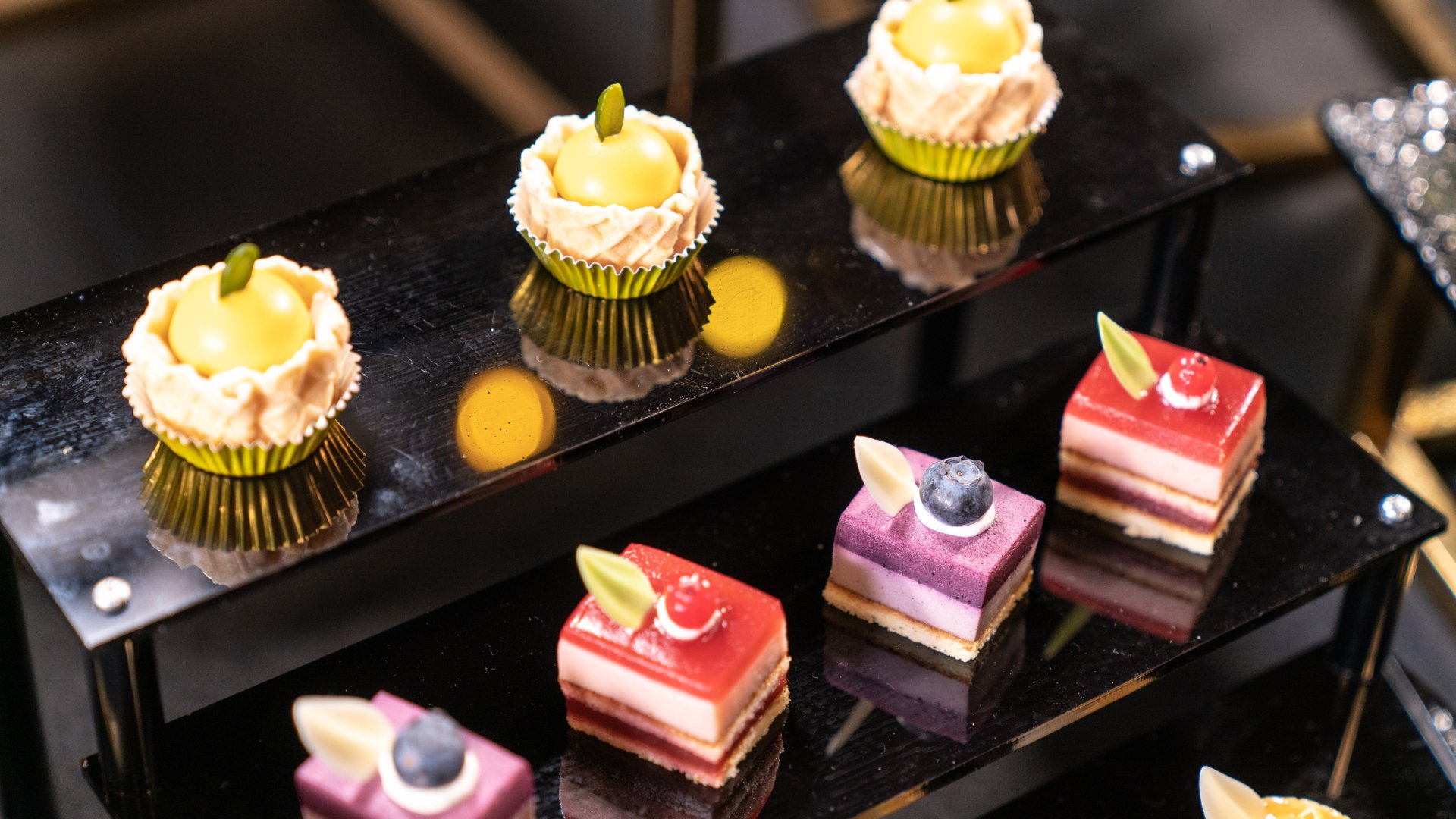 Desserts on black tiered display: Yellow tarts and layered square cakes with berries.