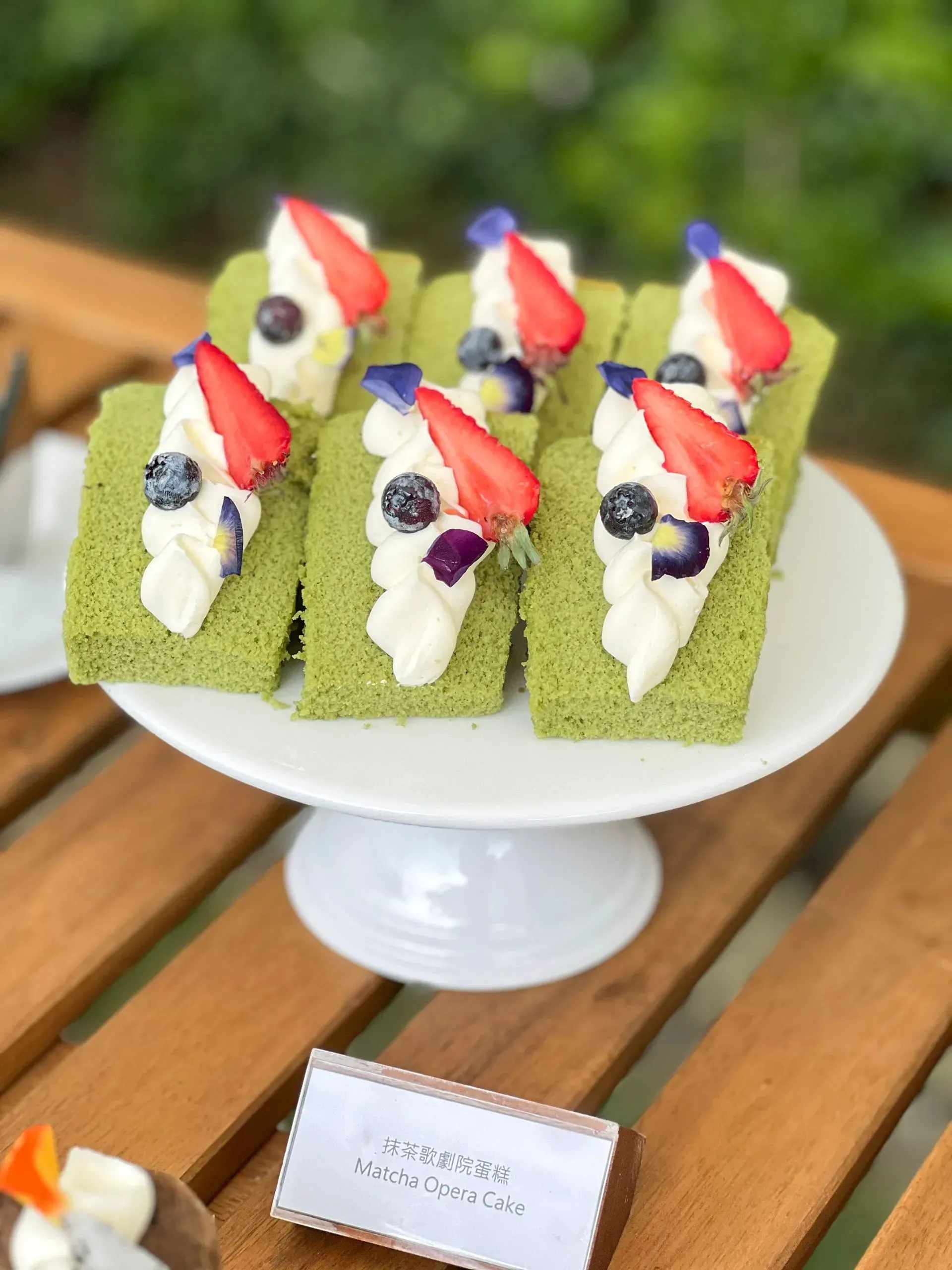 Green matcha cake slices topped with whipped cream, strawberries, and blueberries on a white cake stand.