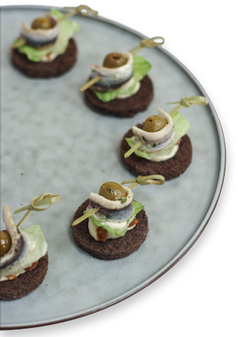 Appetizer bites: round dark crackers topped with lettuce, mayo, anchovy, and olive, skewered on a light blue plate.