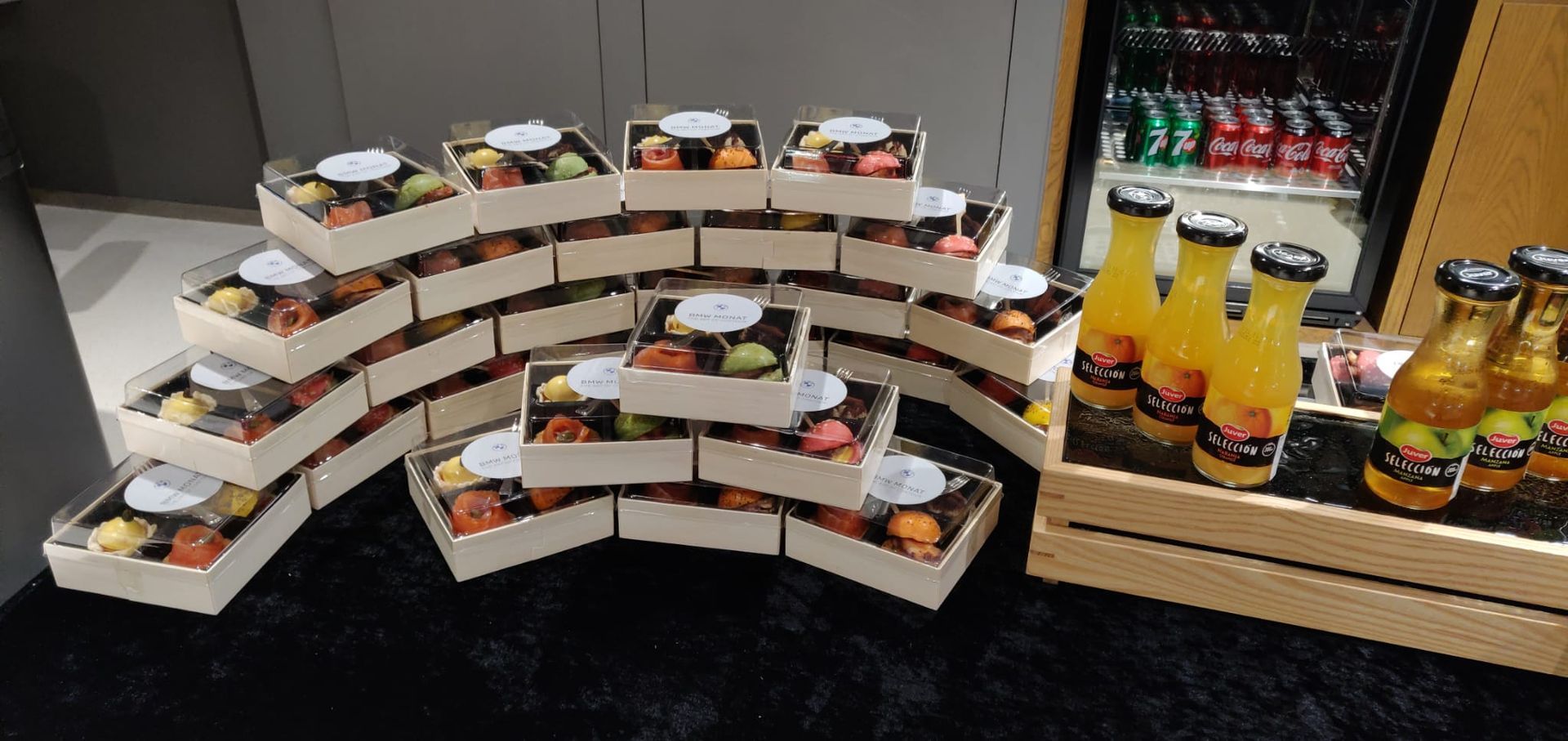 Boxes of colorful macarons are stacked, next to a crate of juice bottles, with a beverage cooler in the background.