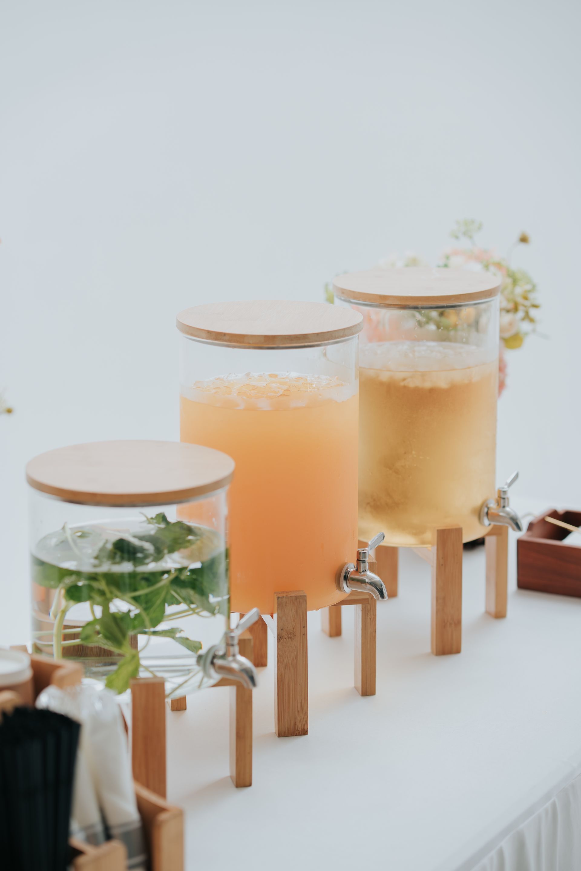 Three beverage dispensers with bamboo lids and stands, containing water with mint, orange juice, and iced tea.