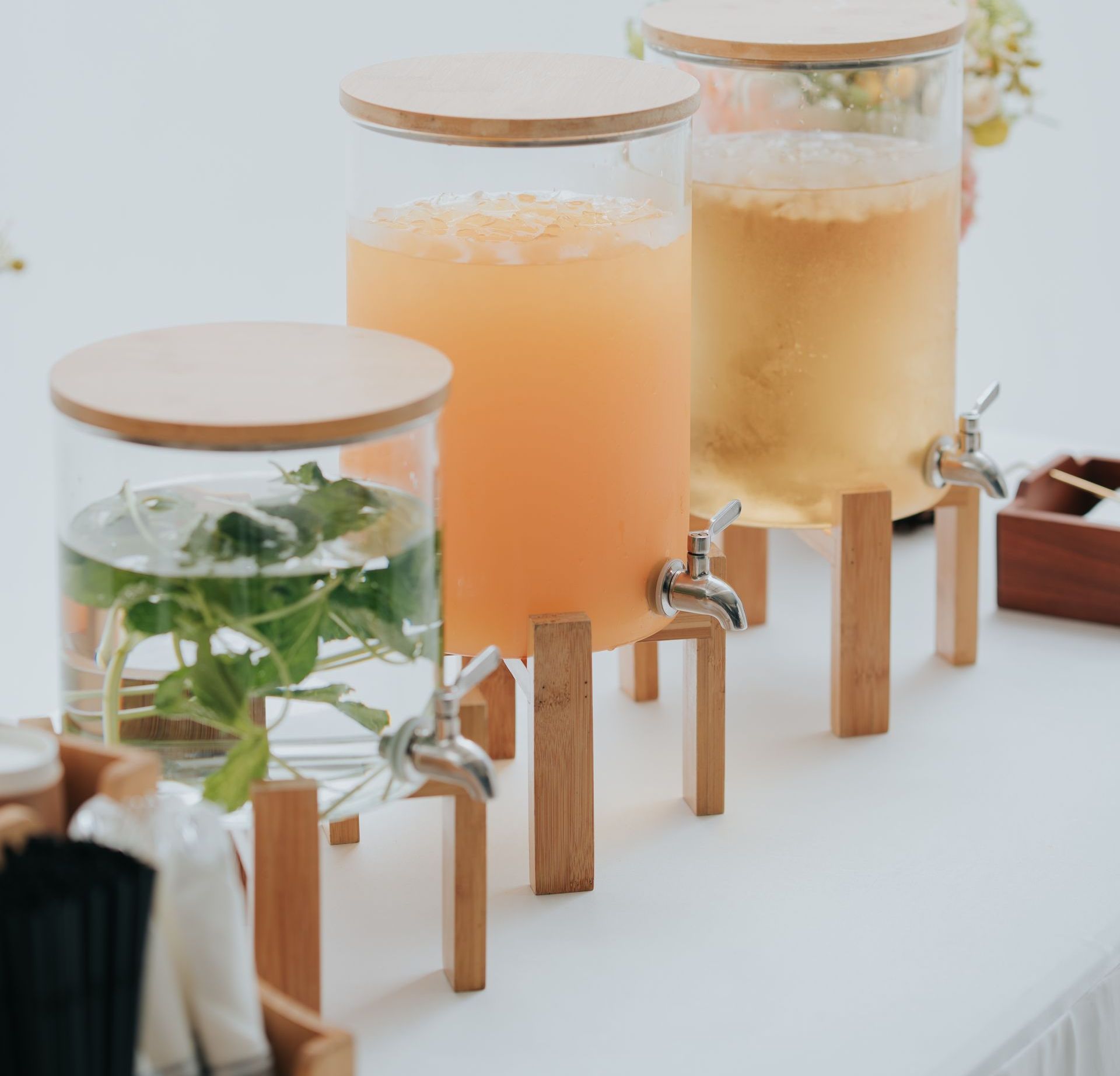 Three beverage dispensers with wooden stands and lids, containing juice and water with herbs, on a white table.