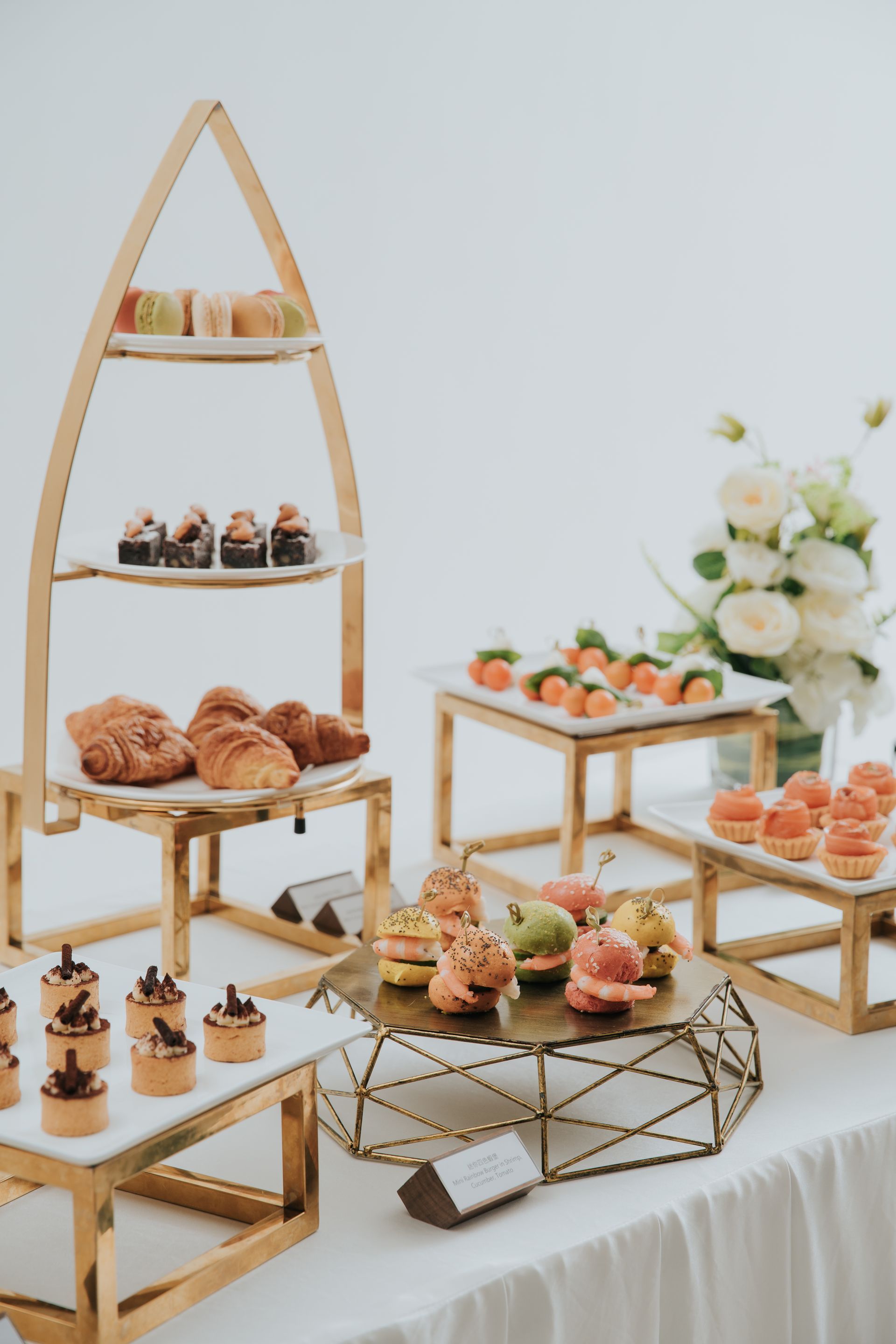 Dessert table with pastries on tiered stands, white tablecloth, gold accents, white background, elegant.