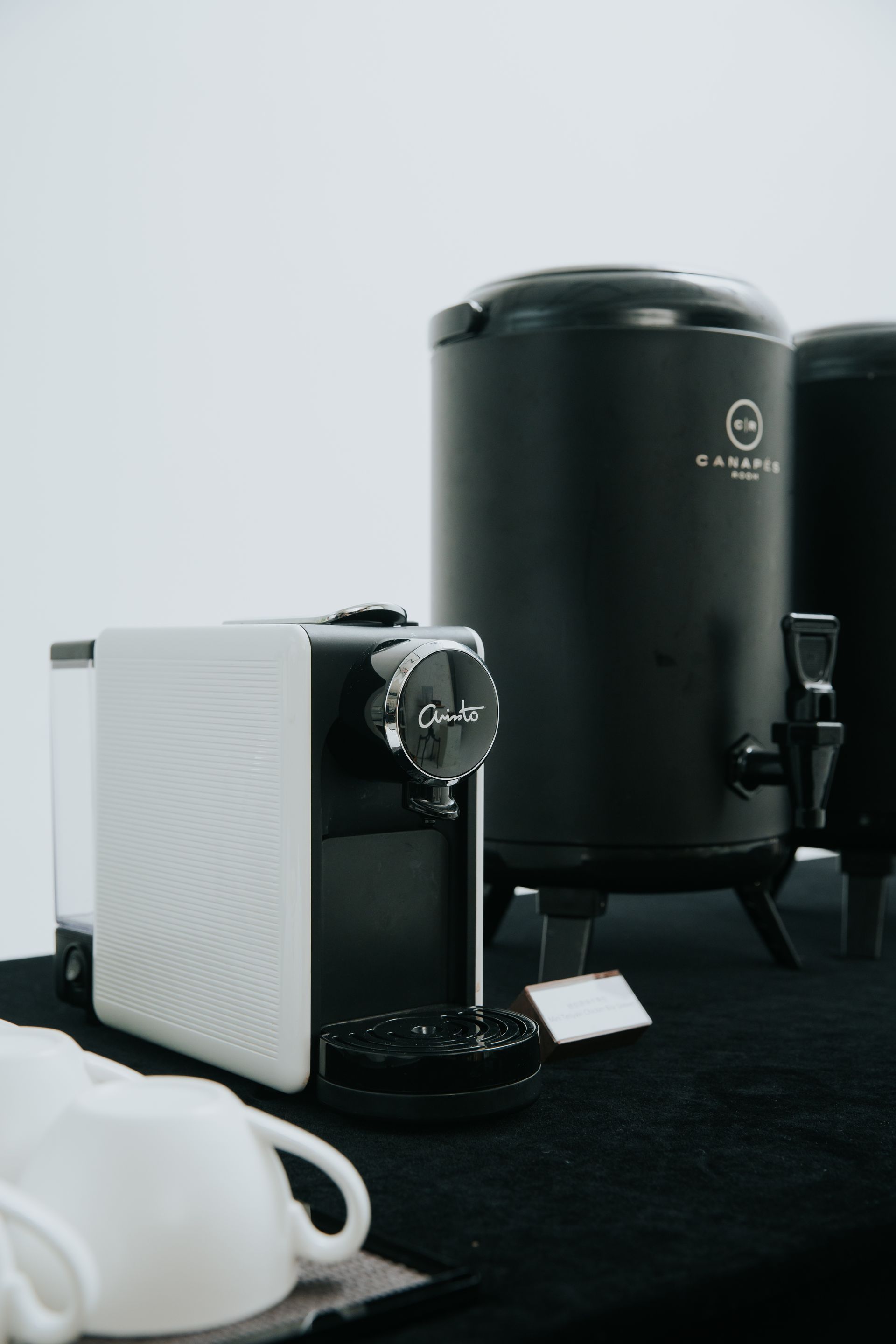 Coffee machine and dispenser on a table; white and black. Cups in the foreground.