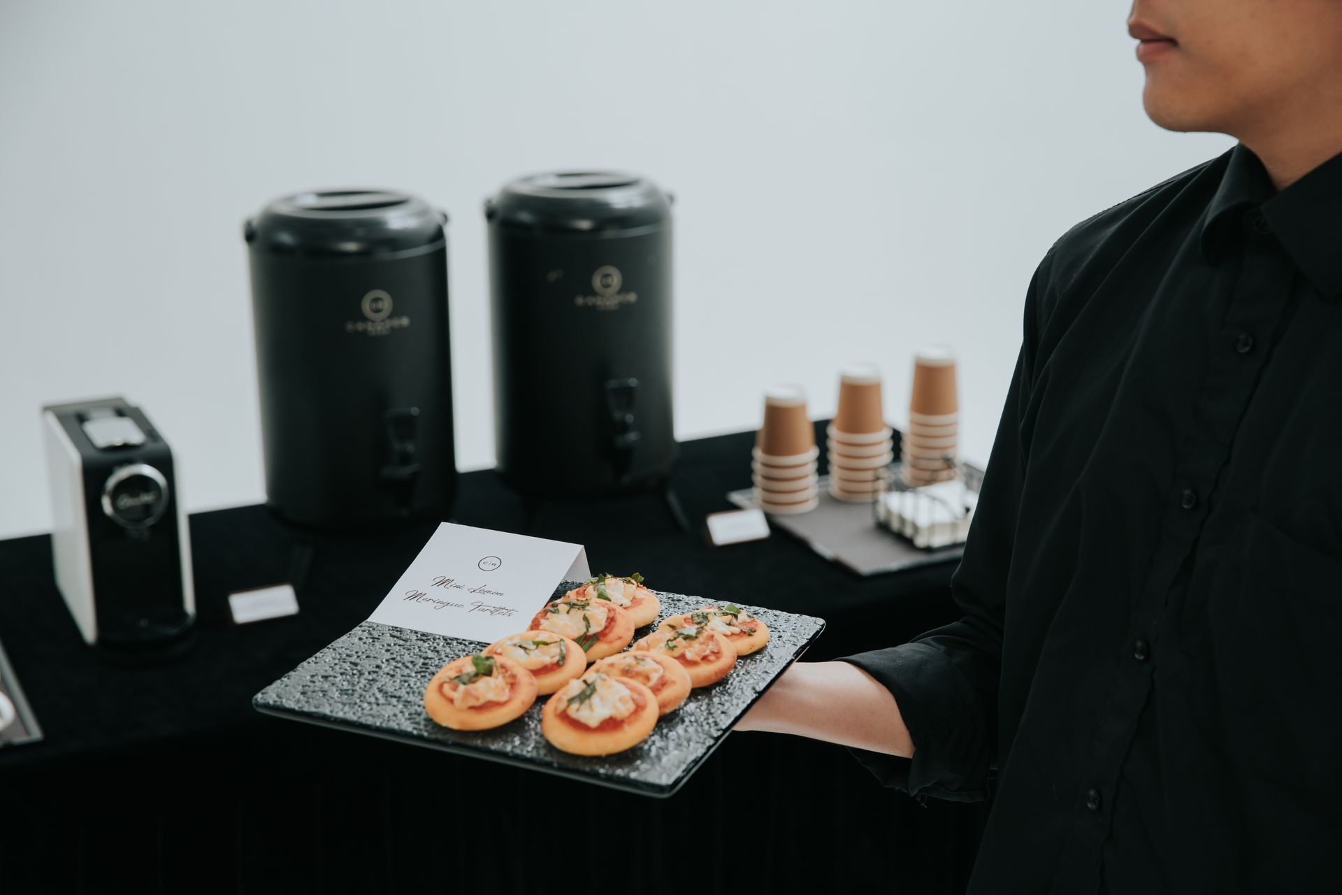 A person holding a plate of appetizers, near coffee makers and cups on a table.