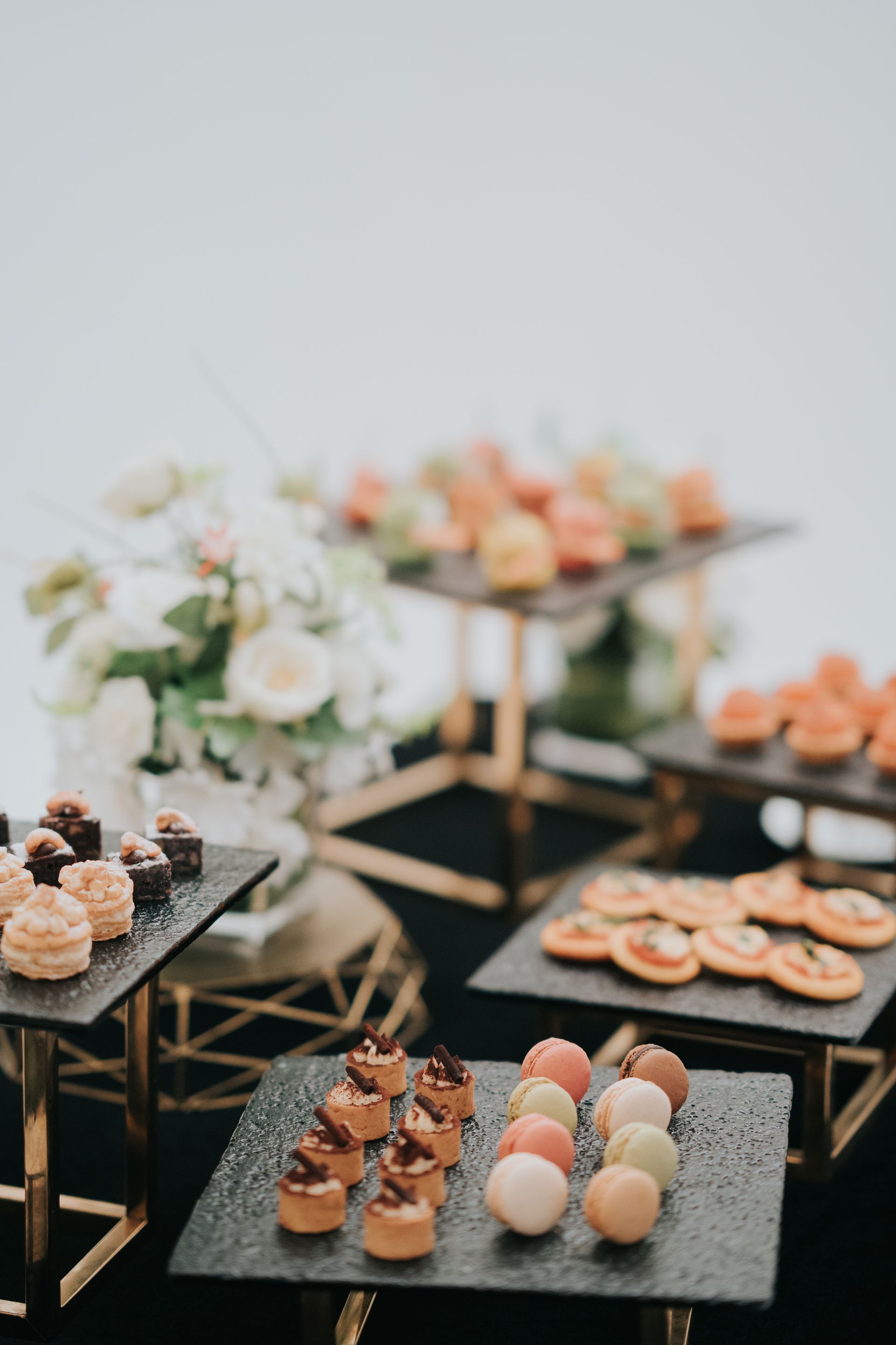 Dessert buffet: assorted pastries on black slate trays with gold stands, white floral arrangement in background.
