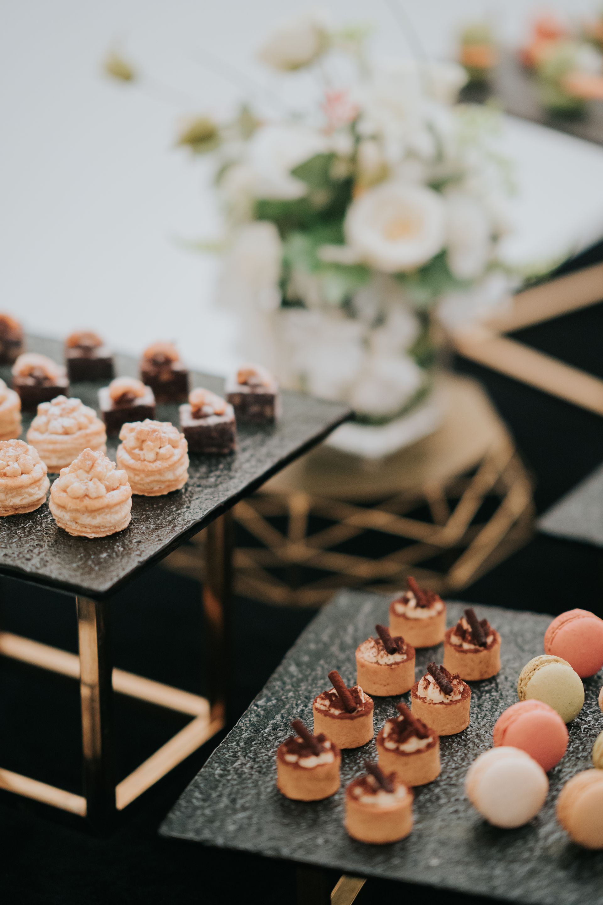 Dessert display: various small pastries and macarons on black serving platters, with flowers in the background.
