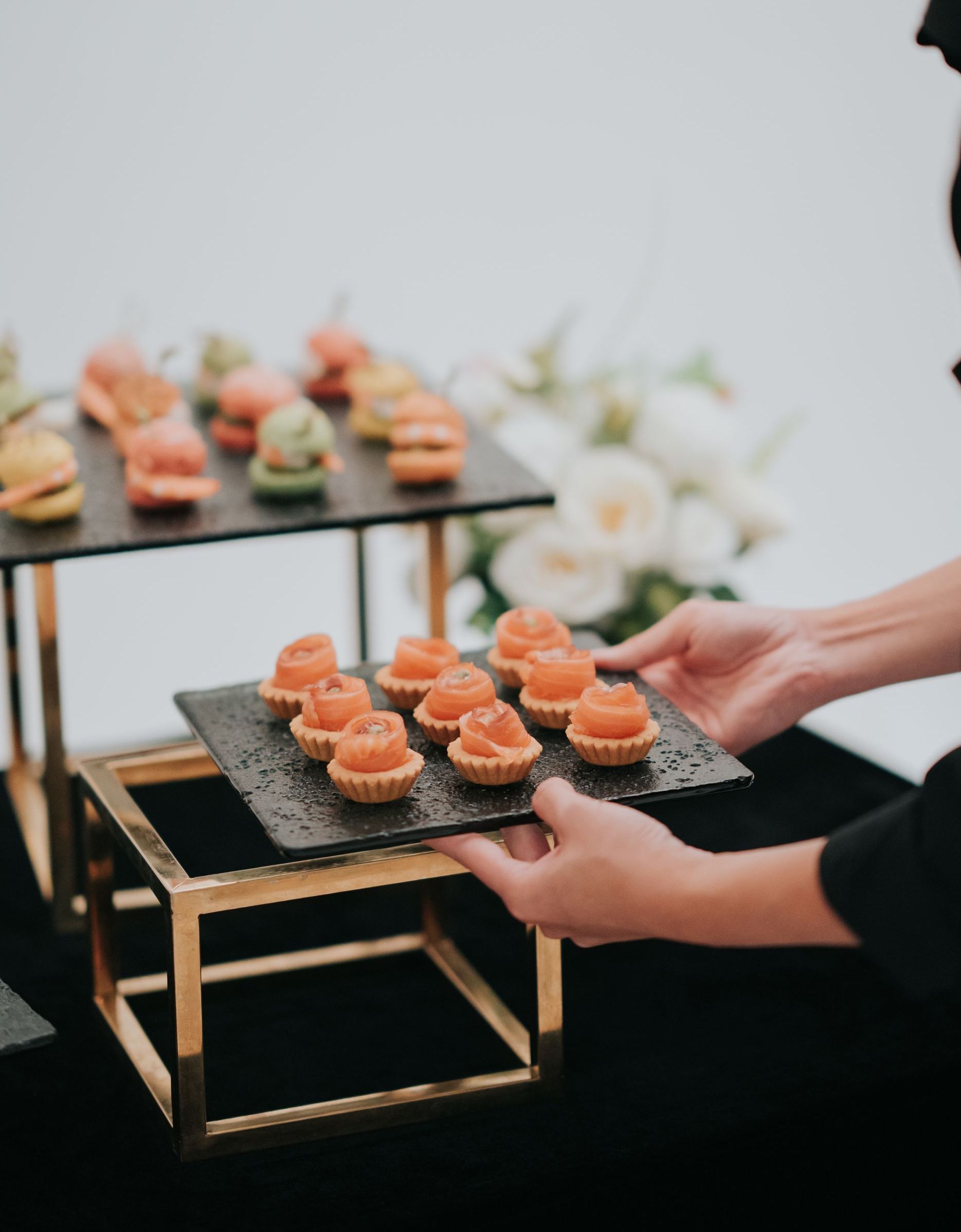 Person arranging hors d'oeuvres on a tiered serving tray with a gold frame and black surfaces; white background.