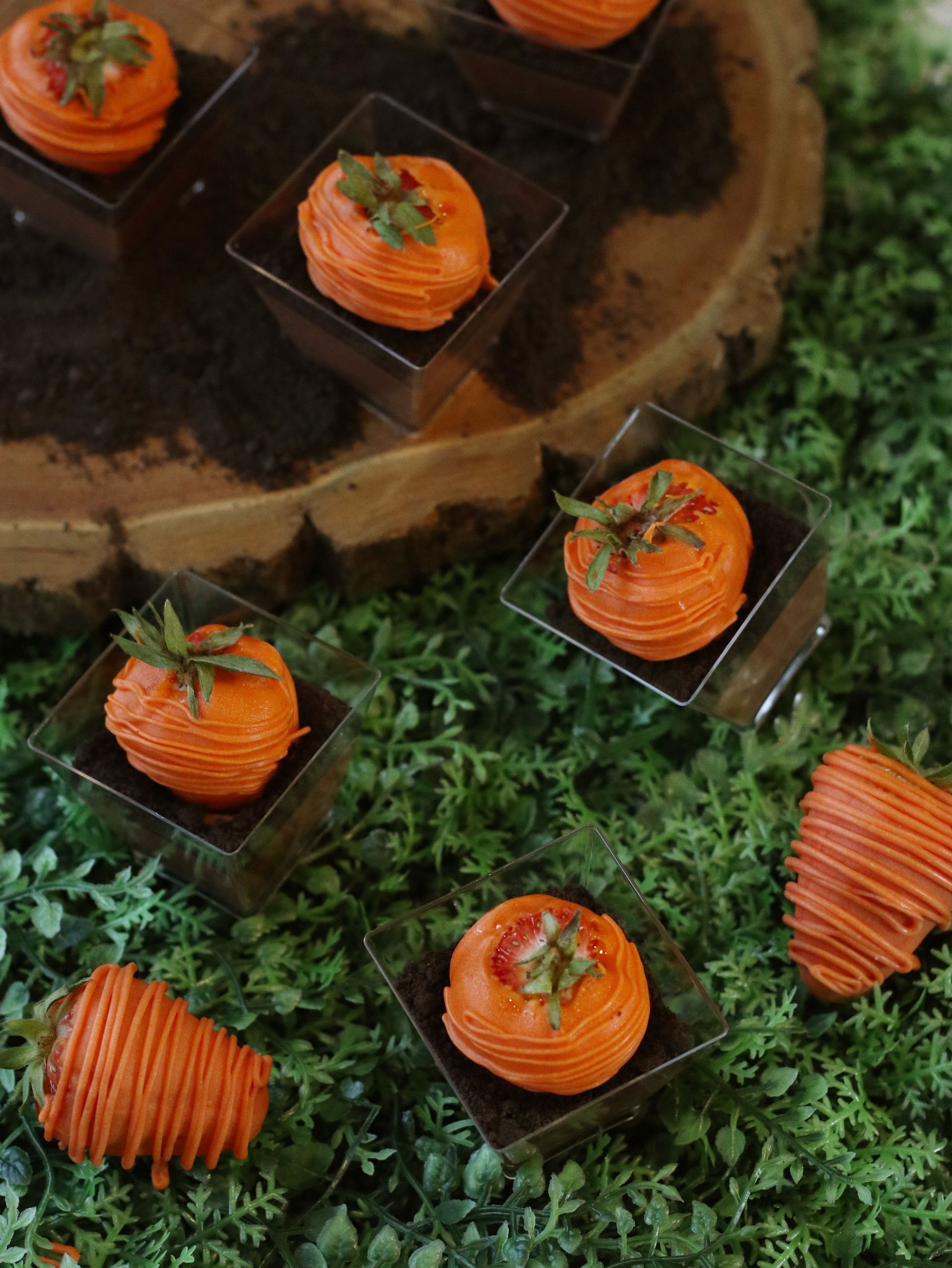 Orange chocolate-covered strawberries in square clear cups, with green garnish, arranged on artificial green grass.