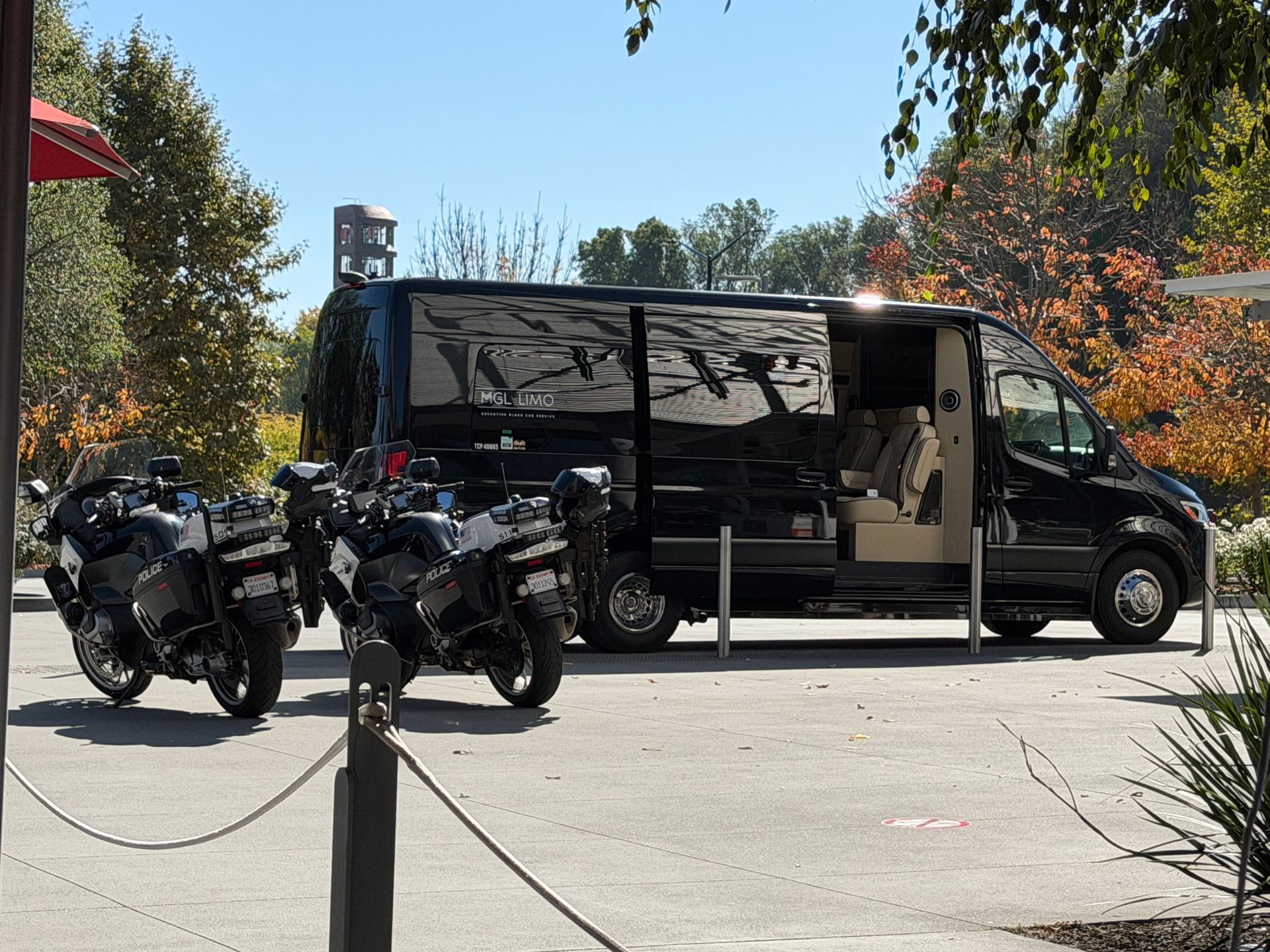 Black van with open side door, two motorcycles parked in front, trees in background.