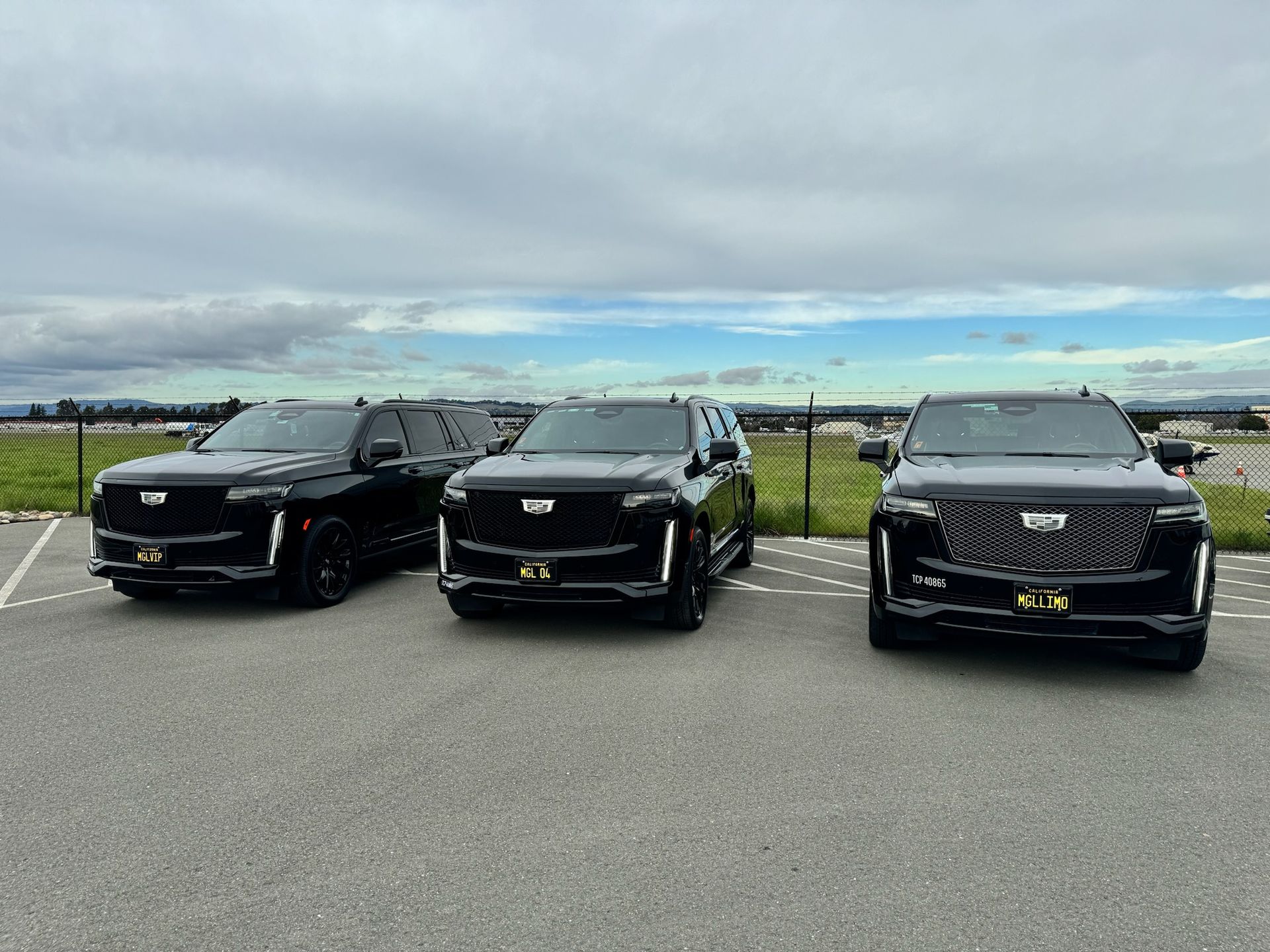 Three black Cadillac Escalade SUVs parked in a lot under a cloudy sky.