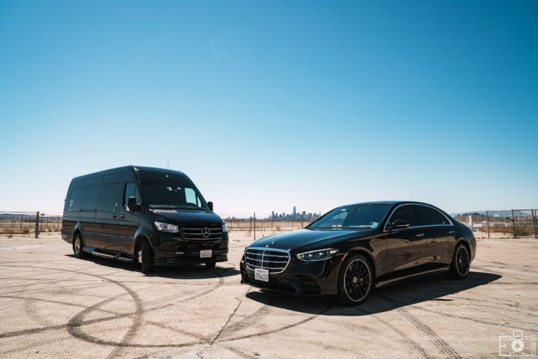 Black van and car parked on dirt, under a clear blue sky. City skyline in the distance.