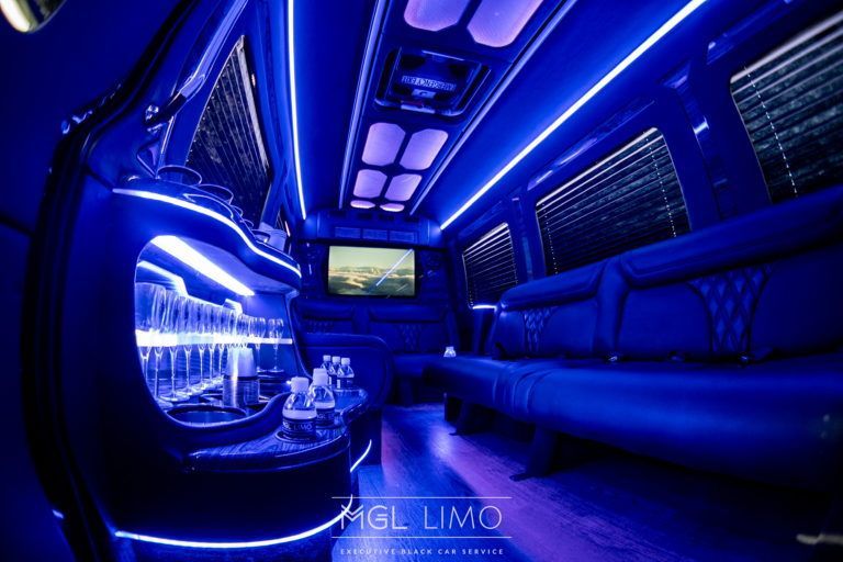 Interior of a limousine with blue neon lights, a bar area with glasses, and a screen.