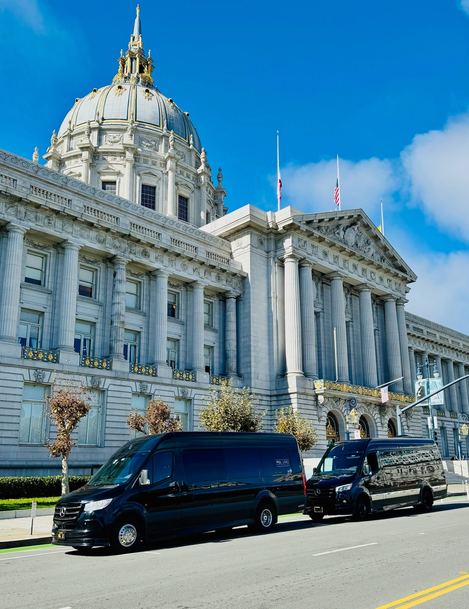 Grand neoclassical civic building with dome and flags on a sunny street, black vans passing in front
