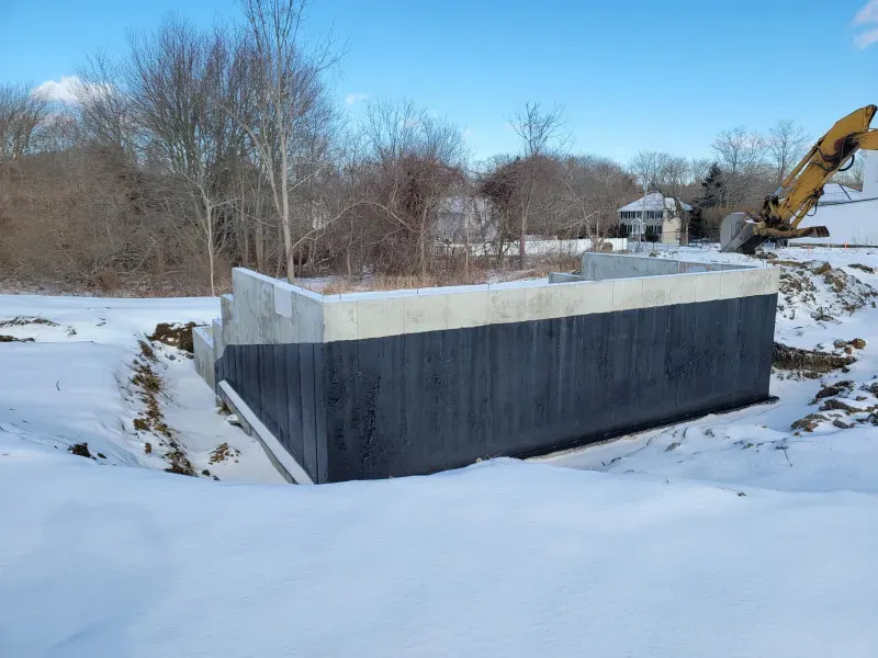 Exterior of concrete foundation coated with black material, surrounded by snow, in a construction site.