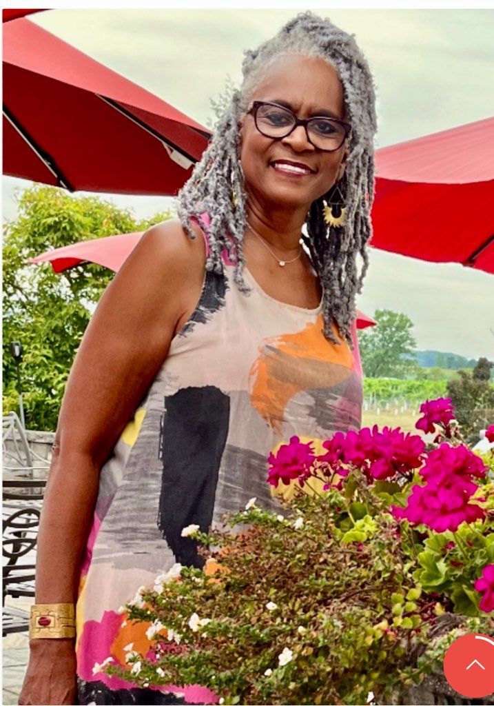 A woman with gray hair and glasses stands in front of flowers and red umbrellas
