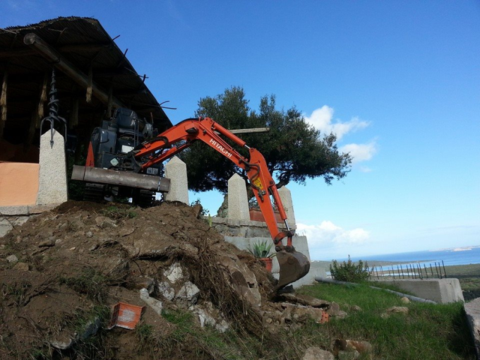Escavatore arancione che scava in un pendio di terra accanto a una casa, su una collina soleggiata con vista sul mare