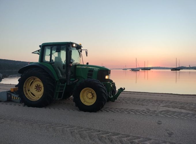 Trattore verde su una spiaggia sabbiosa al tramonto, con il mare calmo e il cielo dai colori pastello.