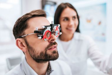 Optometrist adjusting phoropter on man in eye exam room, with assistant in background