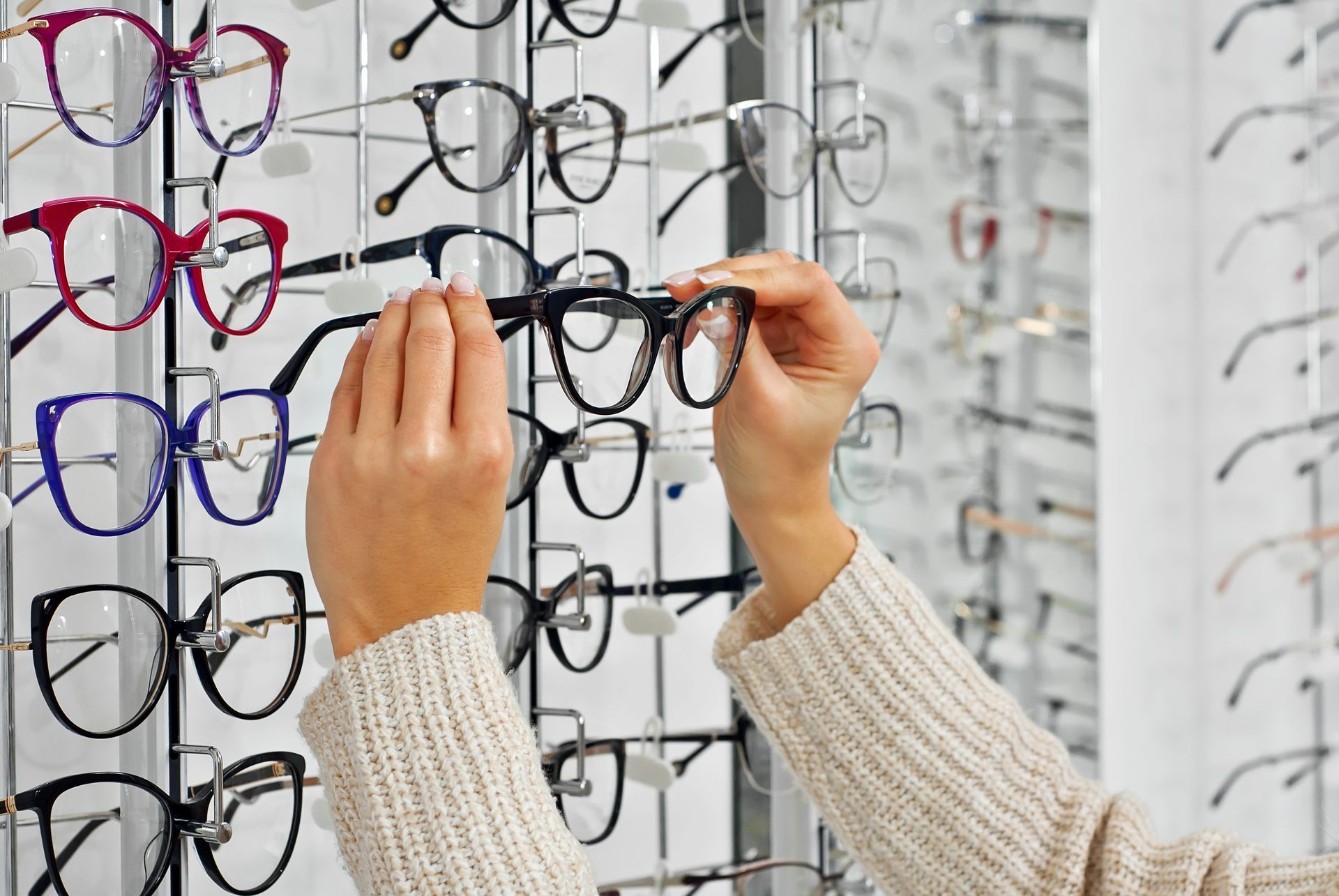 Hands selecting eyeglass frames from a display wall in an optical store