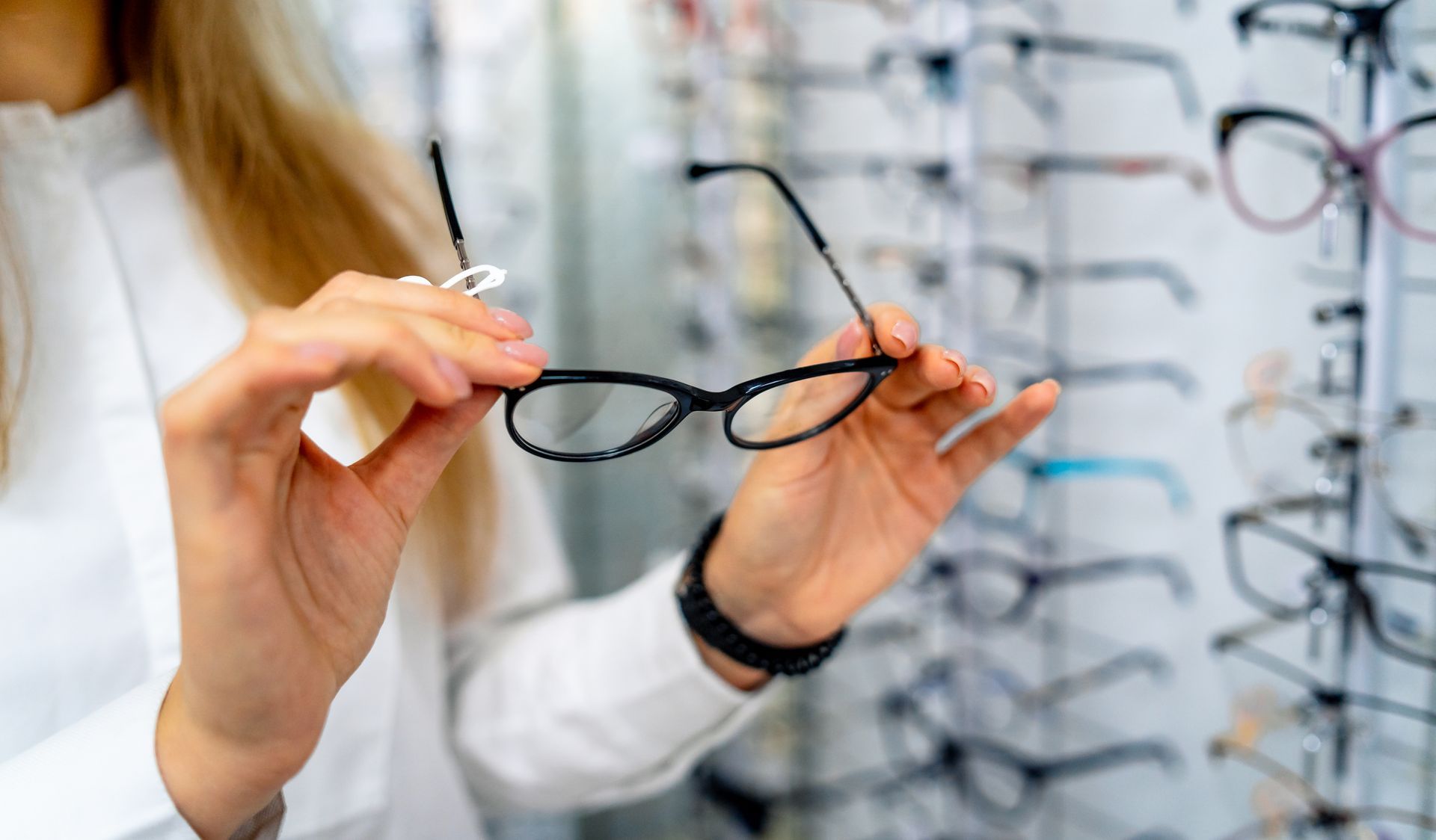 Person holding black eyeglasses in an optical shop, with rows of frames in the background