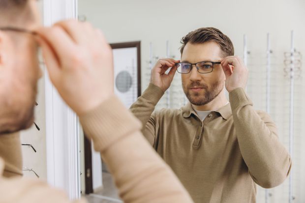 Man adjusting glasses in a bathroom mirror, wearing a tan sweater