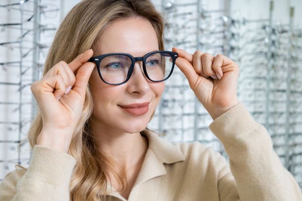 Woman adjusting blue glasses in a bright room with shelves in the background
