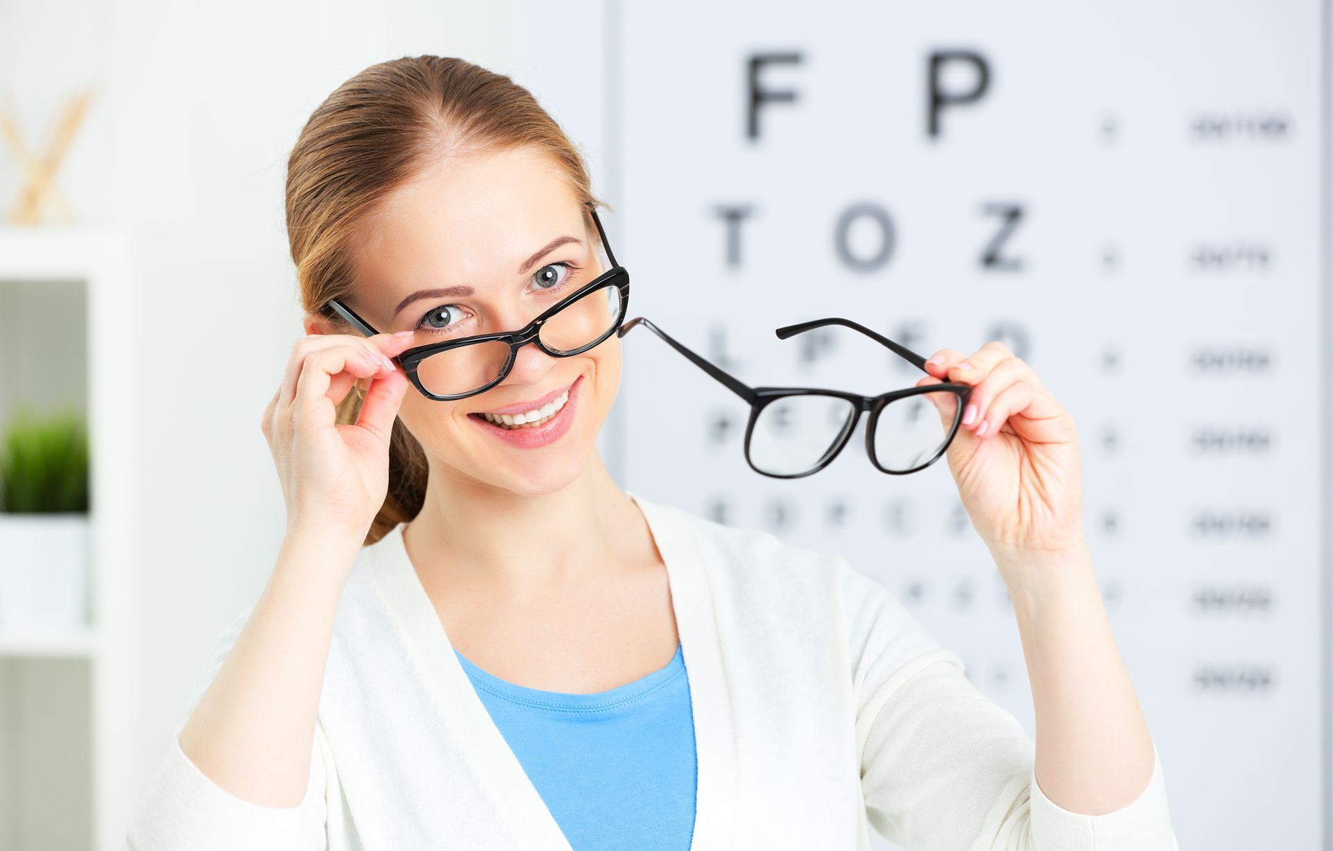 Woman holding eyeglasses in an eye exam room, smiling beside an eye chart